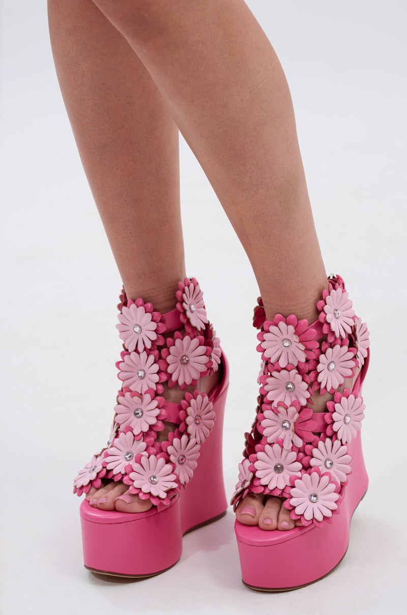 Model showcasing the AZALEA WANG FLOWER EMBELLISHED WEDGE SANDAL IN PINK with multiple large 3D floral appliqués, standing on a white surface.