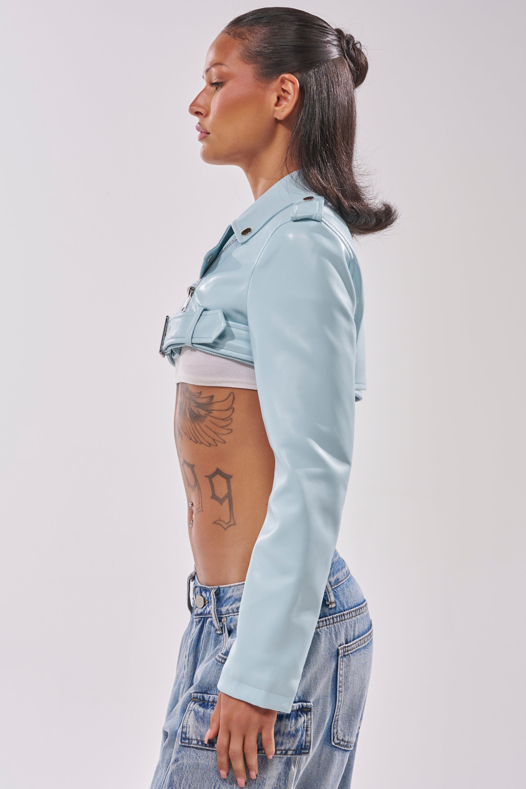 A woman in profile wears the HARLEY LUXE ULTRA CROP MOTO IN LIGHT BLUE with high-waisted jeans. Her slicked-back hair highlights her torso and stomach tattoos against a plain white background. 3