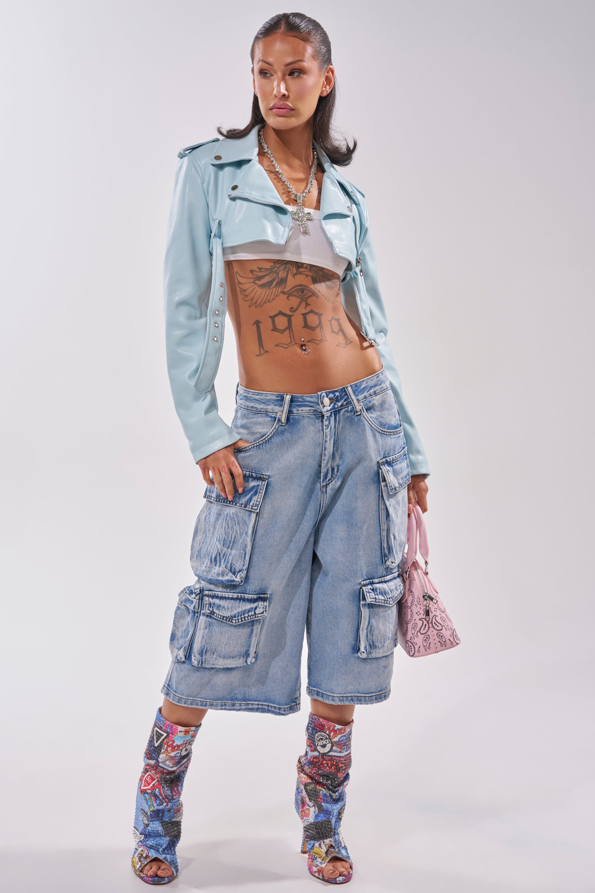 A woman poses in the HARLEY LUXE ULTRA CROP MOTO IN LIGHT BLUE, styled with a white crop top, baggy denim cargo shorts, printed heeled boots, a pink handbag, layered necklaces, and showing her tattooed torso. 6