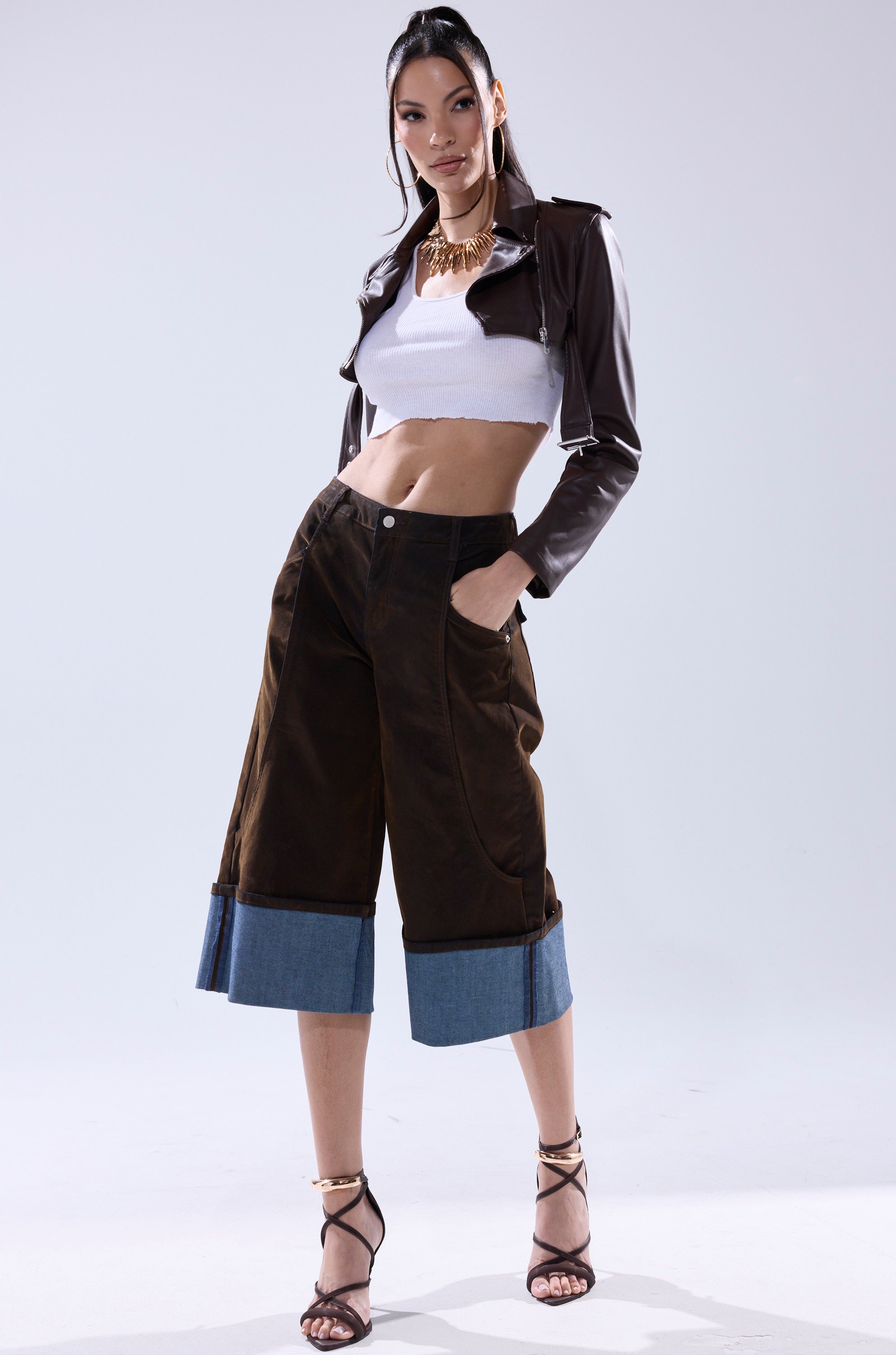 A woman exudes confidence in the HARLEY LUXE ULTRA CROP MOTO IN CHOCOLATE, styled with a white crop top, wide brown pants with denim cuffs, and strappy heels—an effortlessly chic fall look against a light backdrop.