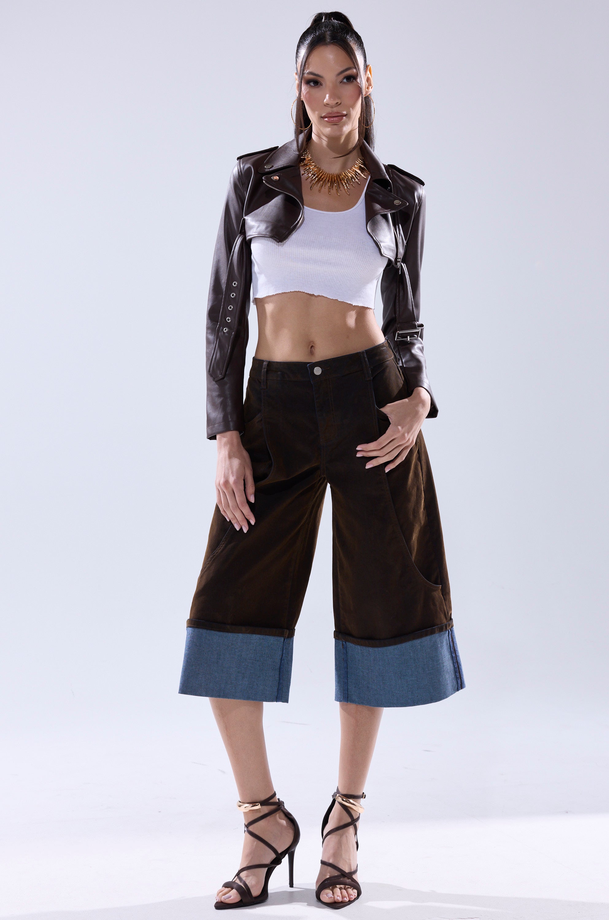 A woman confidently poses in the HARLEY LUXE ULTRA CROP MOTO IN CHOCOLATE, styled with a white crop top, dark wide-leg pants with denim cuffs, black strappy heels, and a statement necklace against a plain light background.