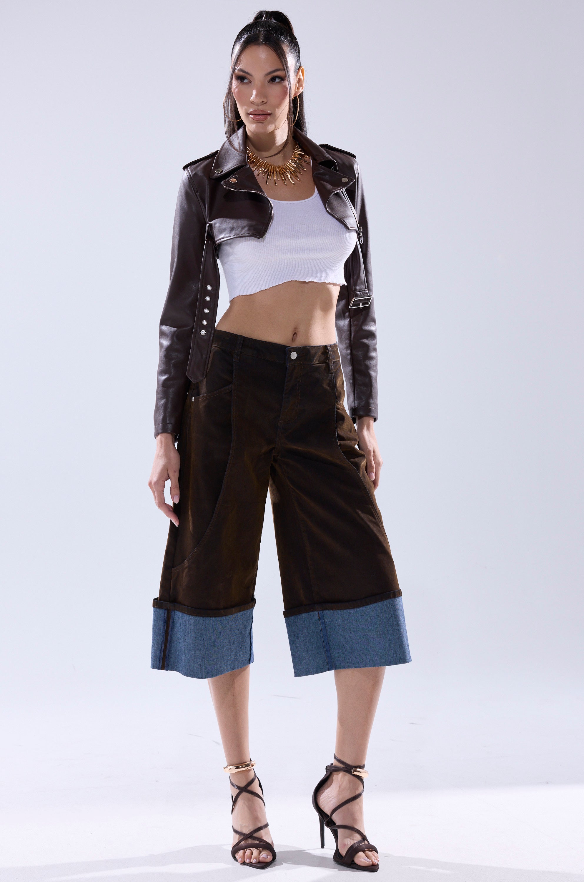 A woman stands confidently in fall fashion, featuring the HARLEY LUXE ULTRA CROP MOTO IN CHOCOLATE jacket, a white crop top, wide-leg dark brown pants with denim cuffs, black high-heeled sandals, and a gold statement necklace.
