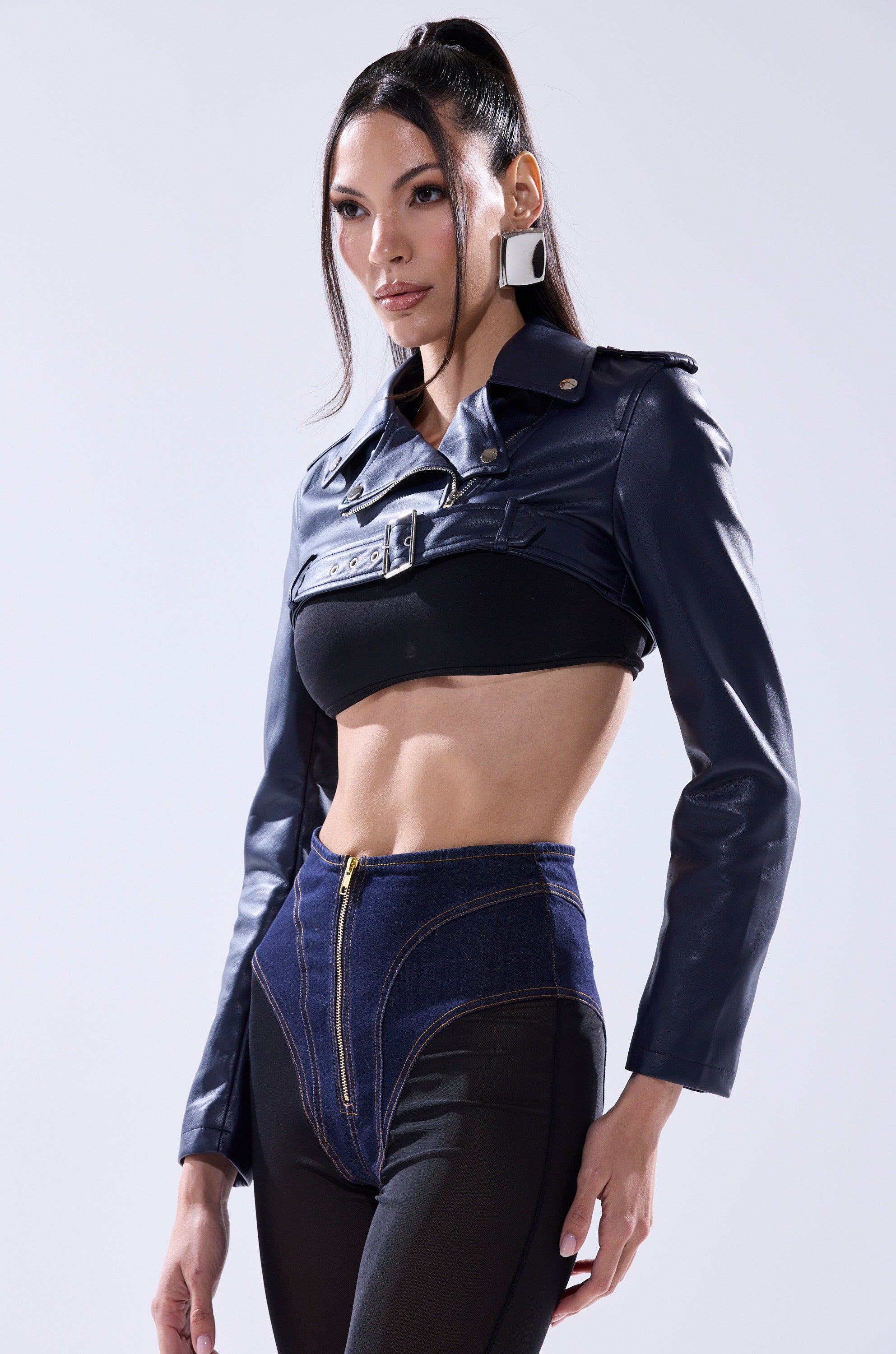 A woman with long dark hair in a high ponytail wears the HARLEY LUXE ULTRA CROP MOTO IN NAVY, a black crop top, high-waisted denim shorts with a gold zipper, and large geometric earrings, posing against a plain light background.