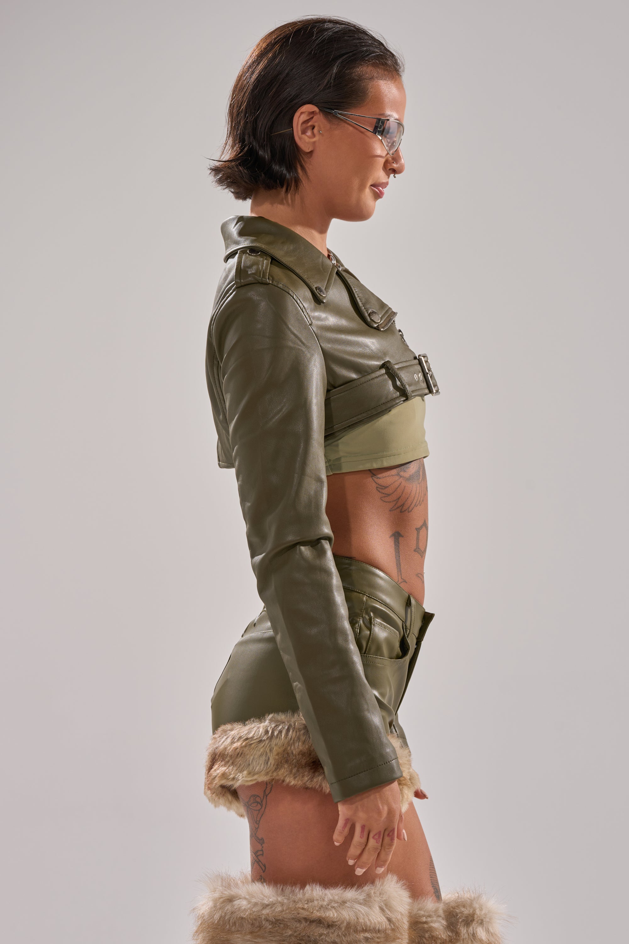 A woman with short dark hair, sunglasses, and tattoos models the HARLEY LUXE ULTRA CROP MOTO IN OLIVE. She pairs it with a matching crop top and fur-trimmed shorts against a plain light background.