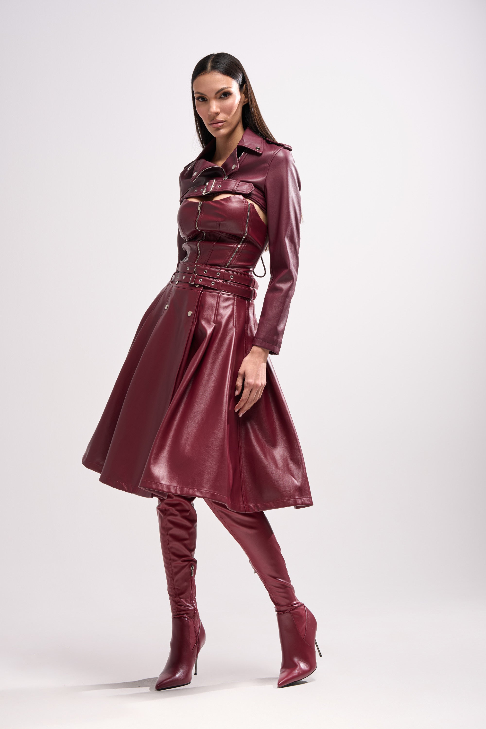 A woman models the HARLEY LUXE ULTRA CROP MOTO IN BURGUNDY, styled over a belted faux leather midi dress with a zipper and long sleeves, creating a chic maroon look perfect for fall and paired with fitted high-heeled boots.