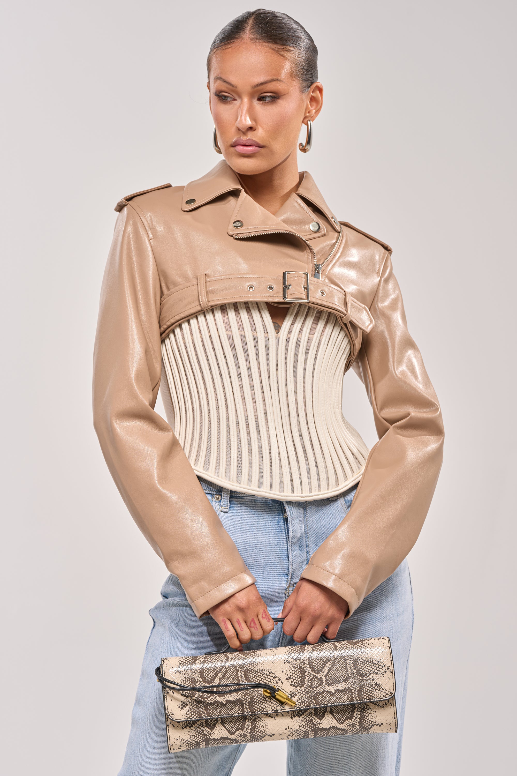 A woman wears the HARLEY LUXE ULTRA CROP MOTO IN TAUPE over a white corset and light blue jeans, accessorized with a snakeskin clutch—an effortlessly chic look for a night out. 0