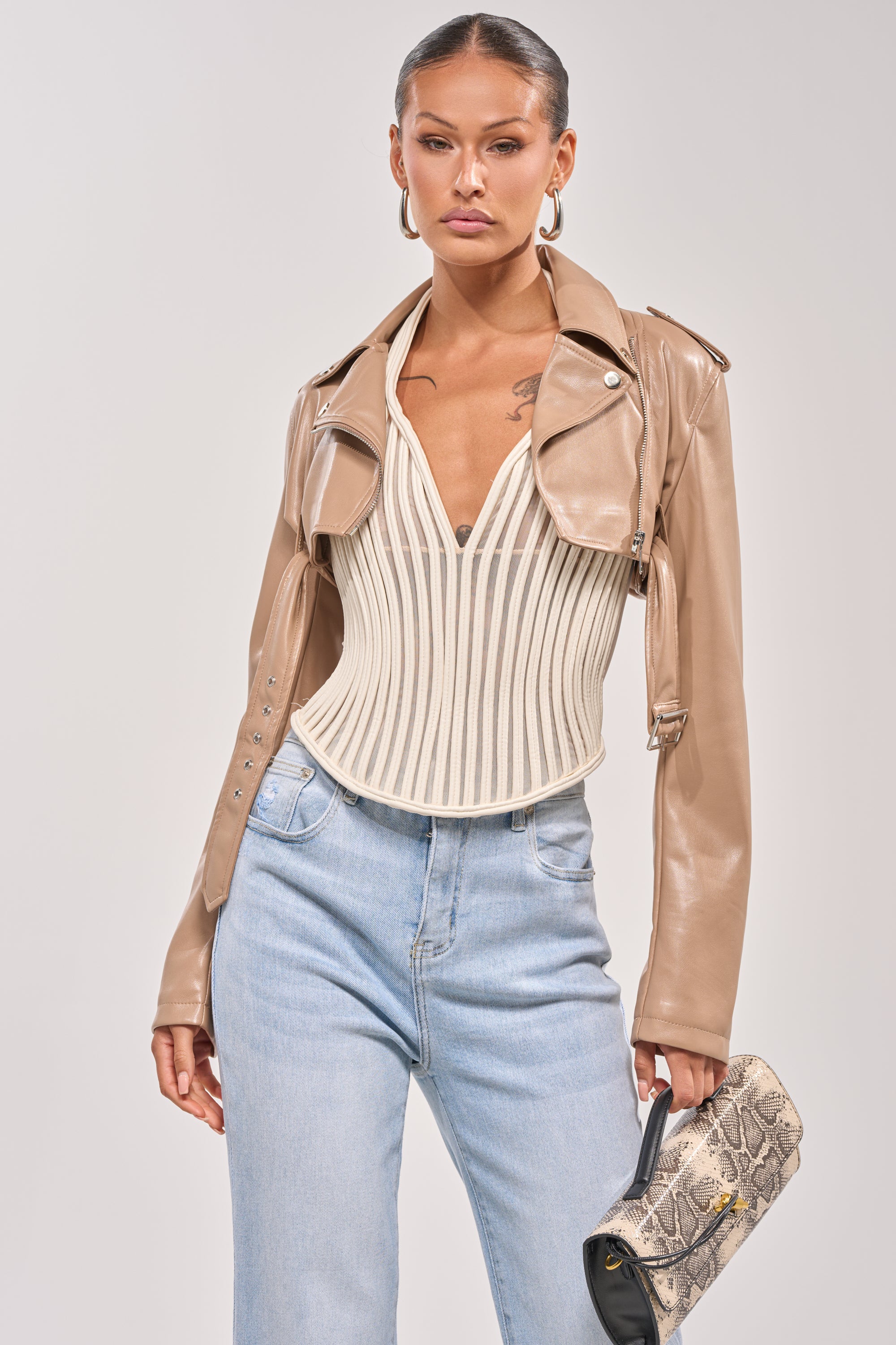 A woman poses in the HARLEY LUXE ULTRA CROP MOTO IN TAUPE, layered over a white corset top with light blue high-waisted jeans and gold hoop earrings, holding a small snakeskin-patterned clutch. 1