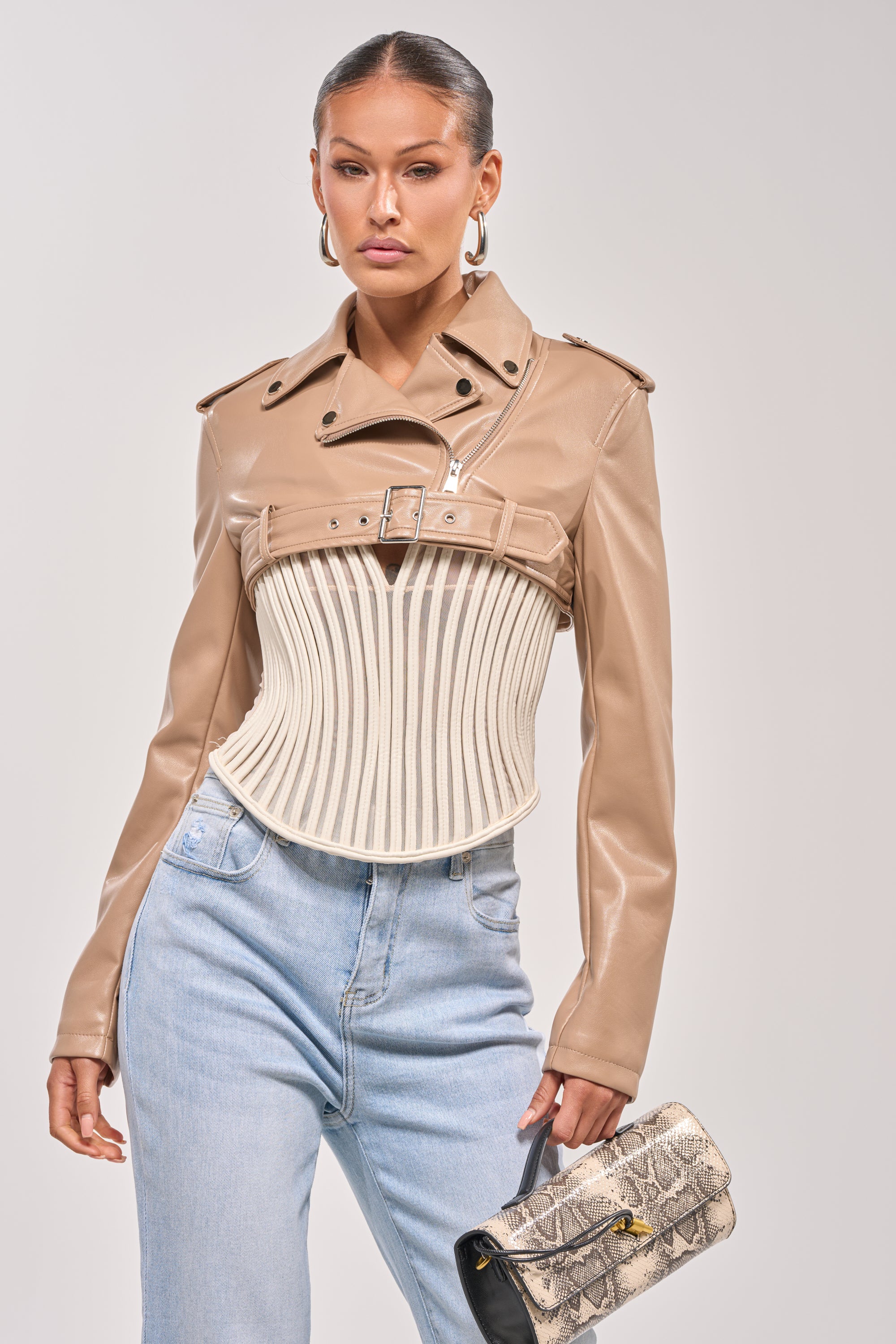 A woman stands against a plain background wearing the HARLEY LUXE ULTRA CROP MOTO IN TAUPE, paired with a structured cream corset detail, light blue high-waisted jeans, large hoop earrings, and holding a snakeskin-patterned clutch bag. 2