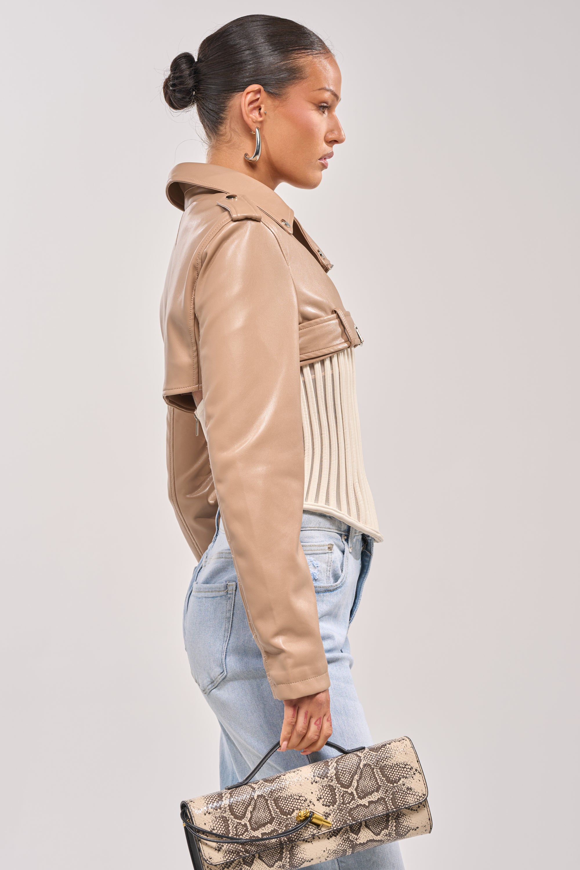 Wearing the HARLEY LUXE ULTRA CROP MOTO IN TAUPE, a woman pairs it with a ribbed cream top, light blue jeans, and a snakeskin clutch. She poses confidently with sleek hair in a bun and large hoop earrings against a plain background. 3