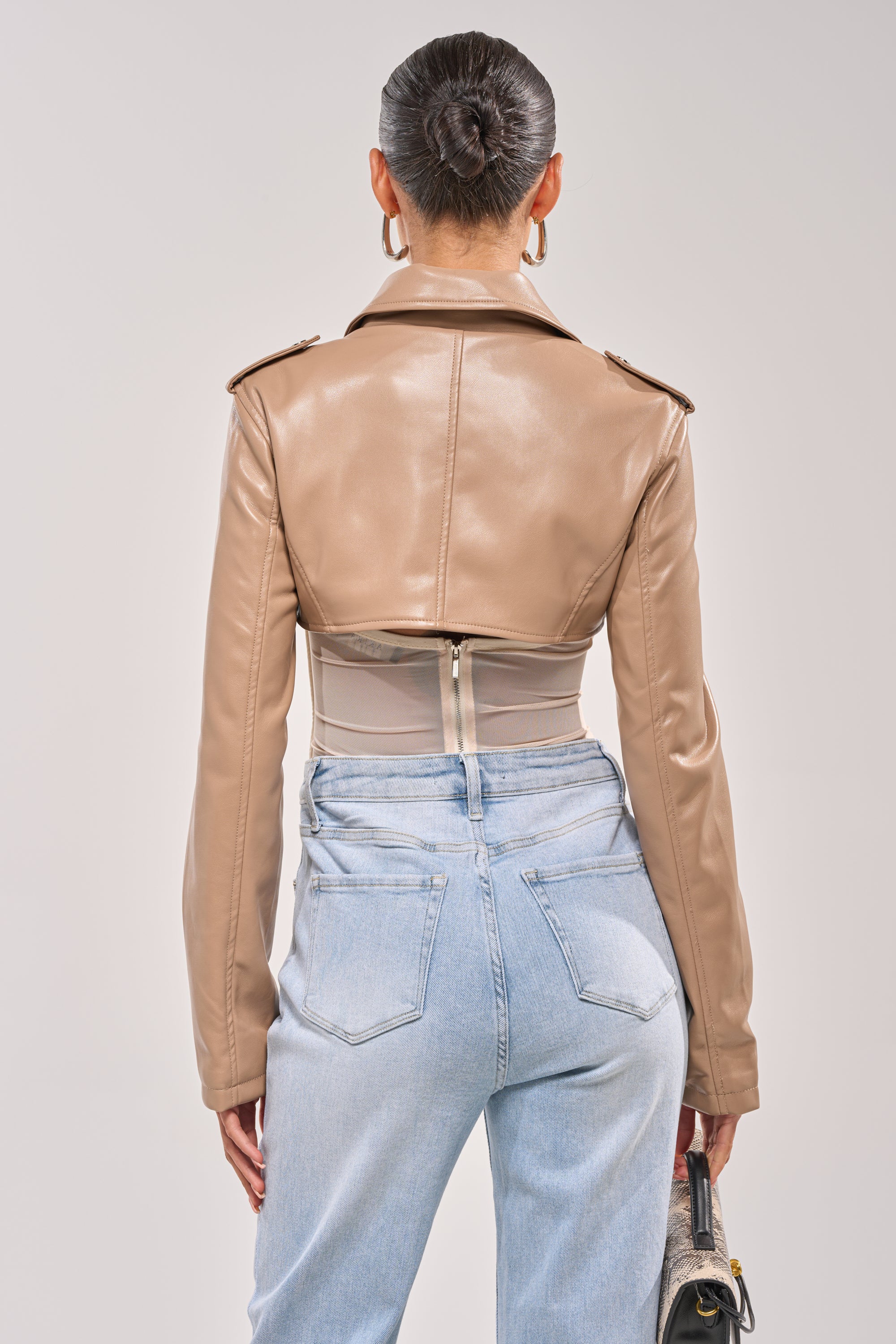 A woman with her hair in a sleek bun and hoop earrings wears the HARLEY LUXE ULTRA CROP MOTO IN TAUPE, light blue high-waisted jeans, and holds a small handbag at her side. 4