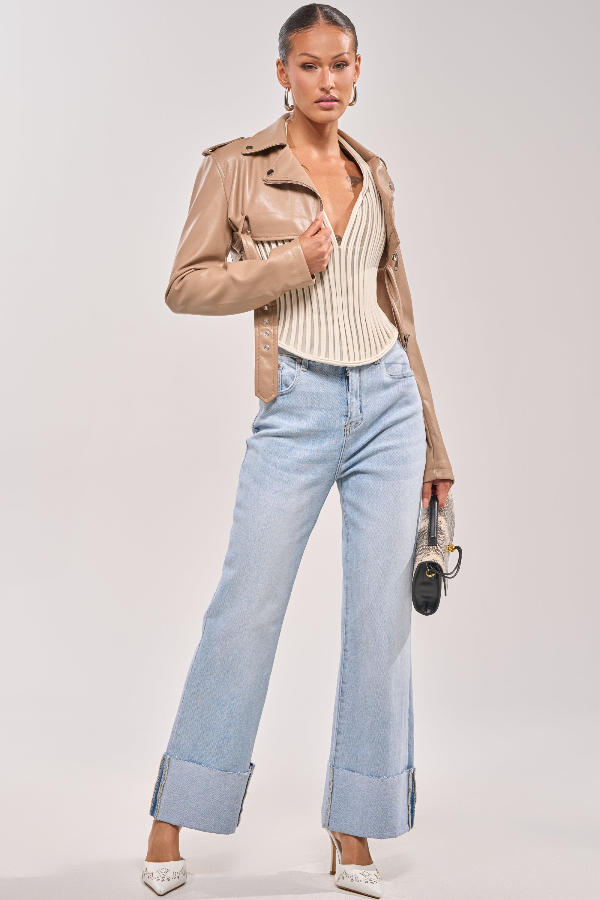 A woman stands confidently in the HARLEY LUXE ULTRA CROP MOTO IN TAUPE over a cream ribbed V-neck top, light blue wide-leg cuffed jeans, white heels, and a small handbag against a plain light background. 6