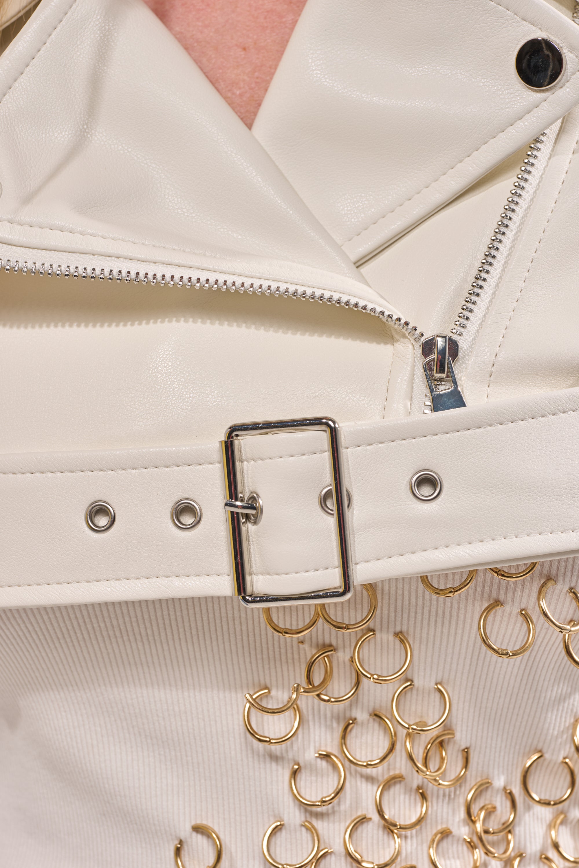 Close-up of the HARLEY LUXE ULTRA CROP MOTO IN WHITE, a cropped white faux leather jacket with a silver zipper, belt buckle, and gold ring embellishments below the belt. 8