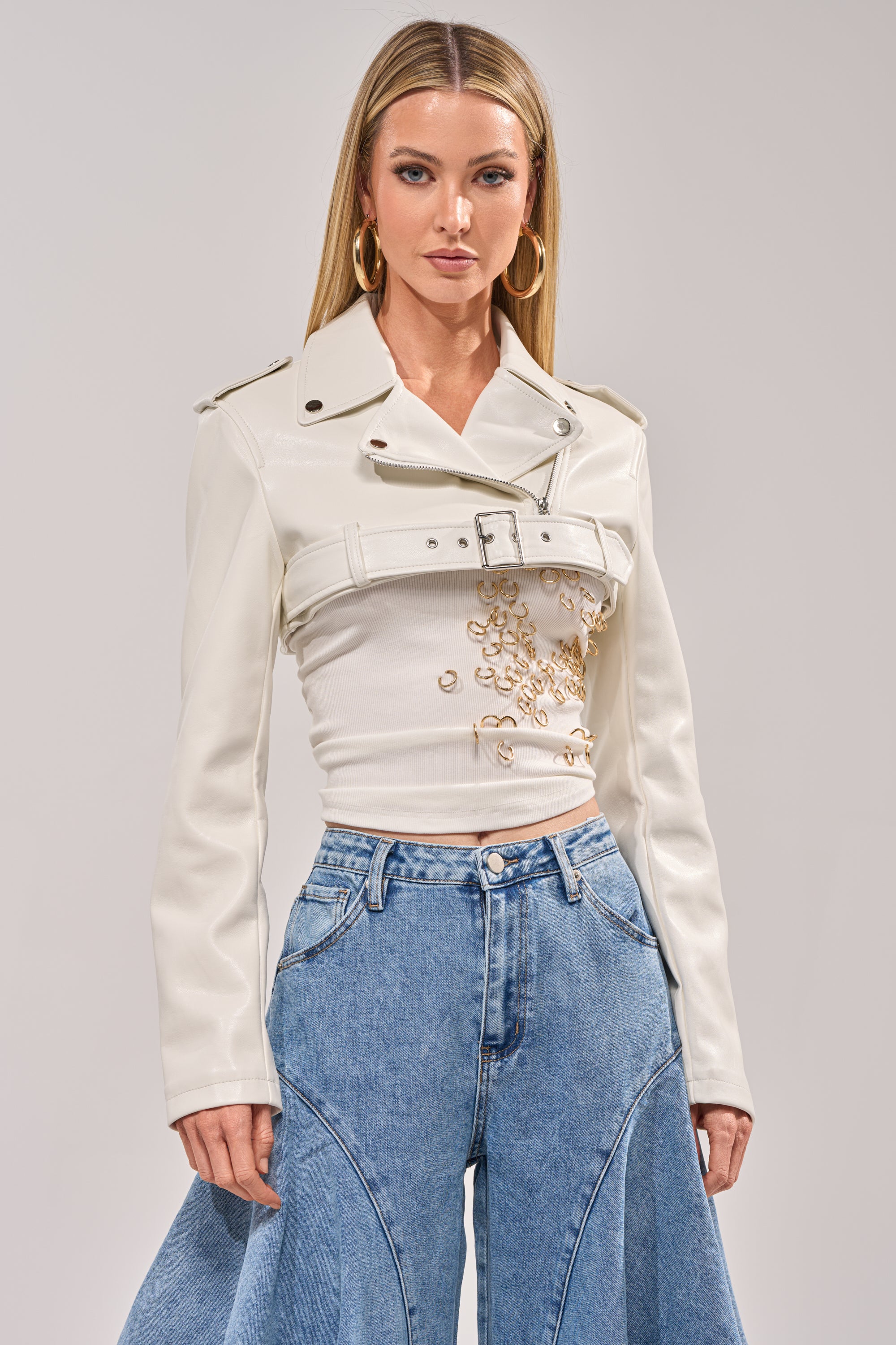 A woman with long, straight blonde hair and large hoop earrings faces forward, wearing the HARLEY LUXE ULTRA CROP MOTO IN WHITE over a detailed white top and high-waisted blue jeans with unique seam designs. 2