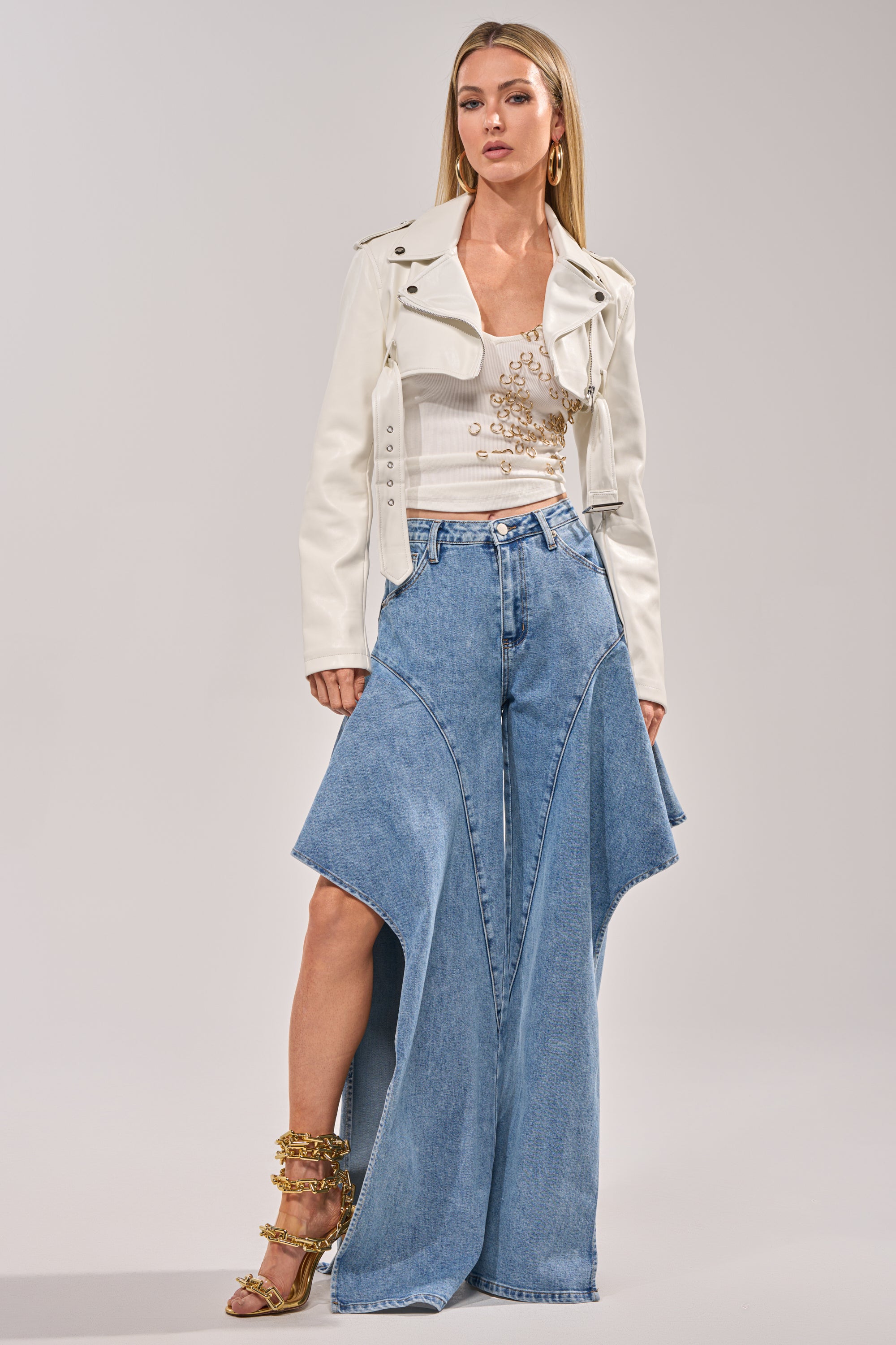 A woman poses confidently against a plain background in the HARLEY LUXE ULTRA CROP MOTO IN WHITE, paired with a white chain-detail top, wide-leg blue jeans with flared panels, gold strappy heels, and long straight hair. 6