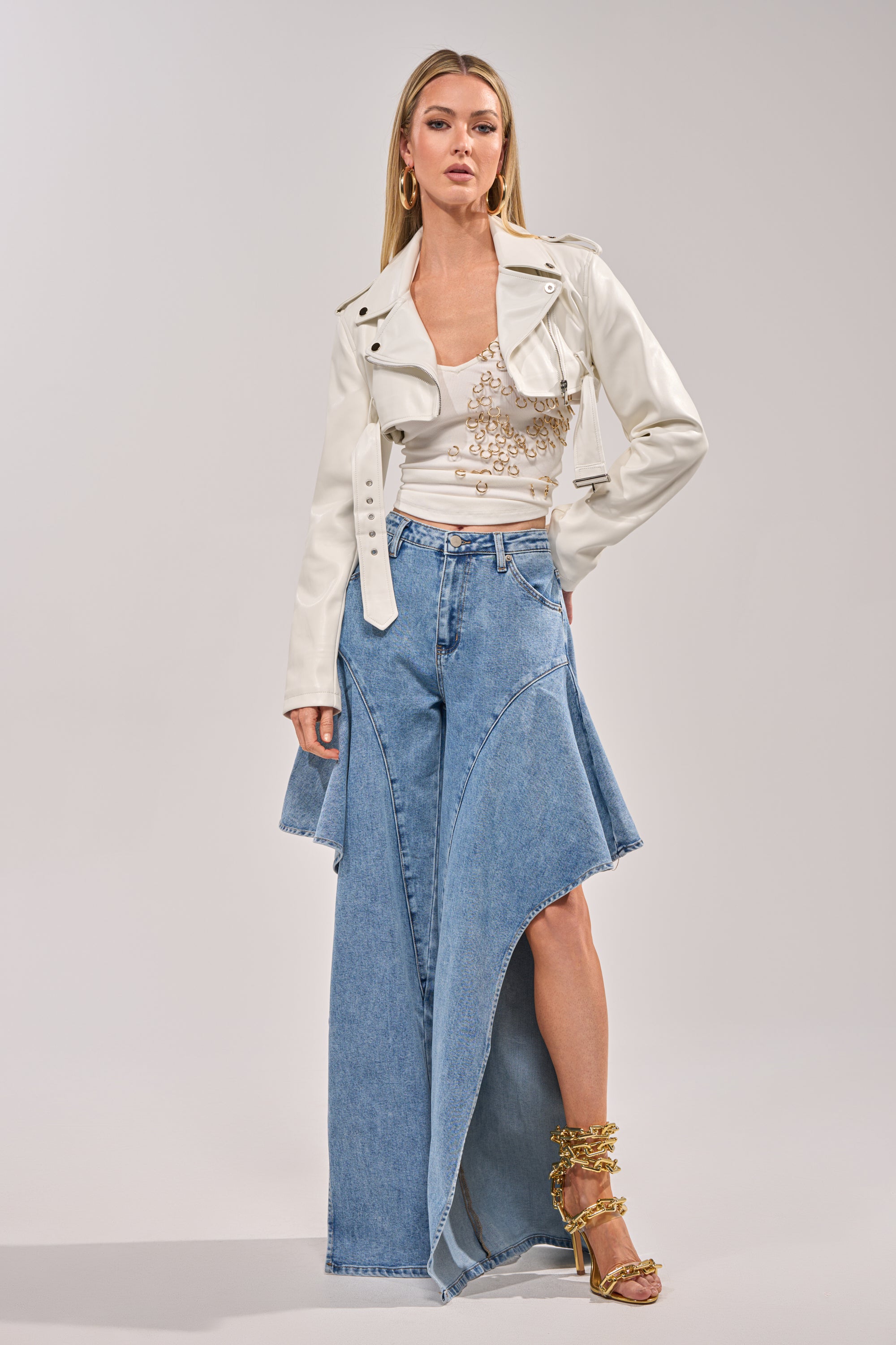 A woman poses in the HARLEY LUXE ULTRA CROP MOTO IN WHITE faux leather jacket, a beaded white top, and a blue asymmetrical denim maxi skirt with a high slit. She pairs gold strappy heels and hoop earrings against a light gray background. 7