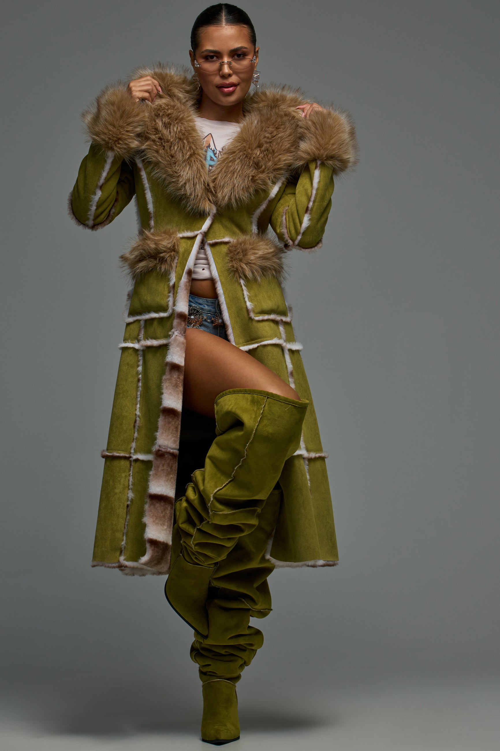 Wearing the JUNEAU FAUX SUEDE COAT with faux fur trim, a woman stands confidently against a plain gray background. She pairs the olive coat with matching high boots, glasses, hoop earrings, and a white shirt.