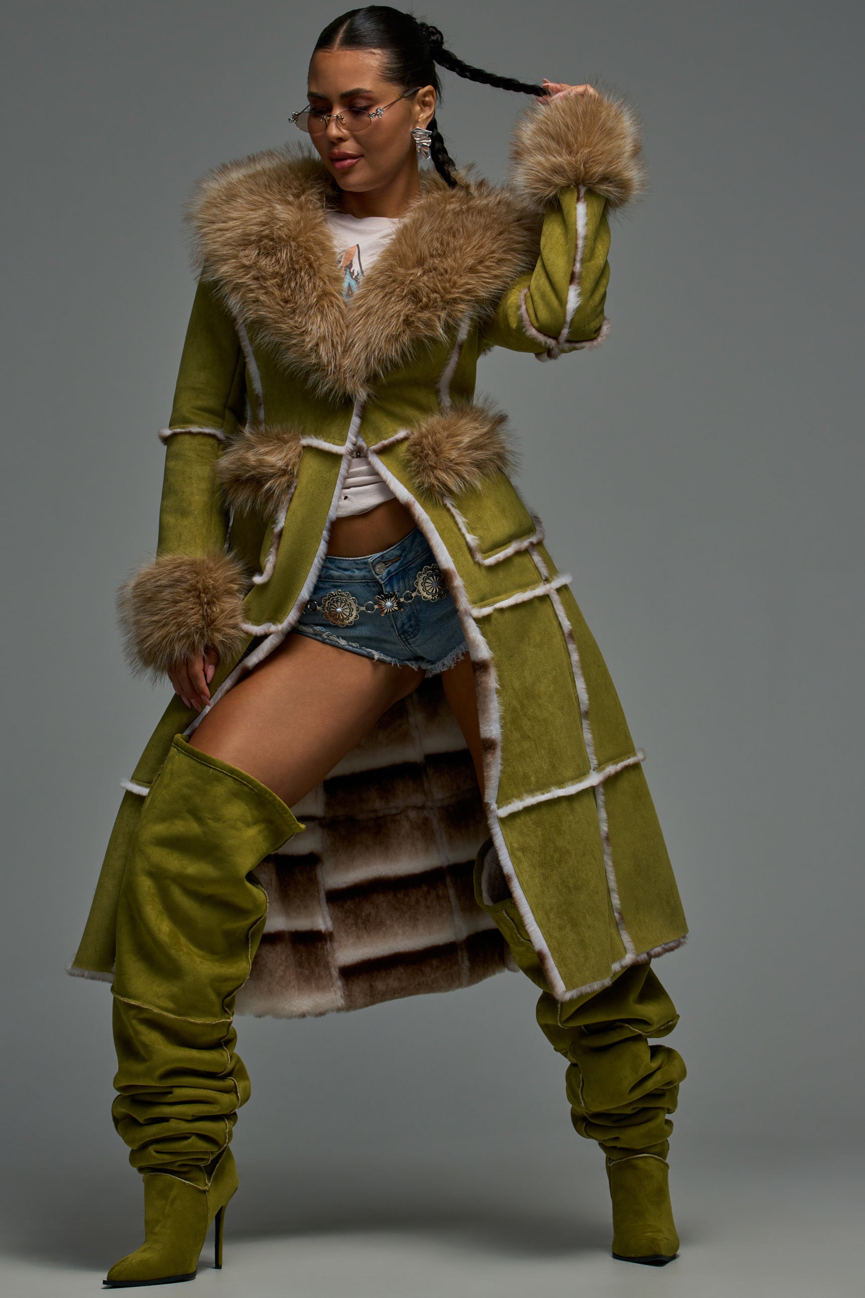Woman poses in the HARLOW FAUX SUEDE FUR TRENCH COAT IN OLIVE, paired with denim shorts, thigh-high green boots, and sunglasses against a gray backdrop for a chic longline look.