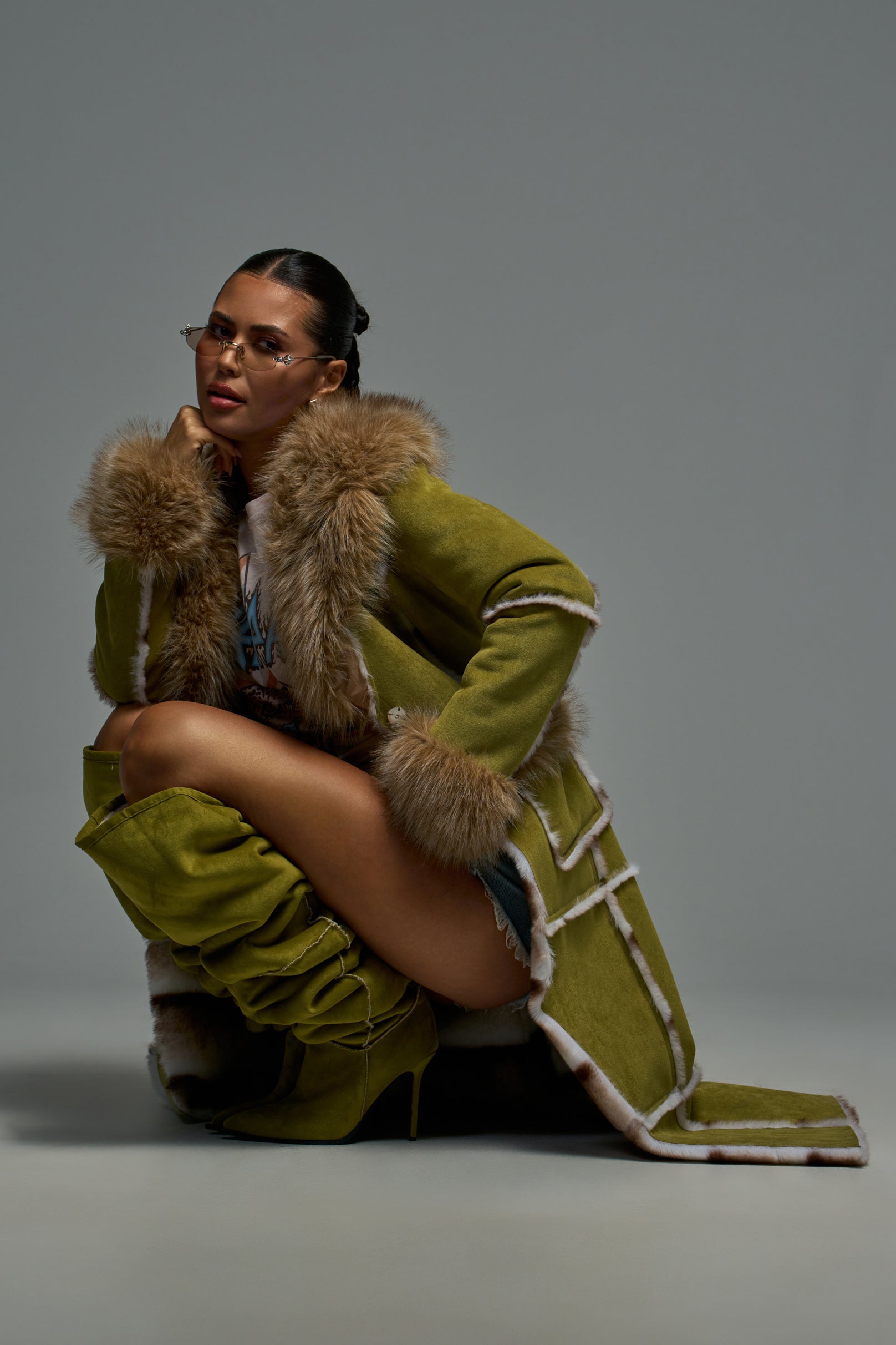 A woman poses confidently in the HARLOW FAUX SUEDE FUR TRENCH COAT IN OLIVE, styled with matching boots, glasses, and denim shorts against a plain gray background for a chic longline look.