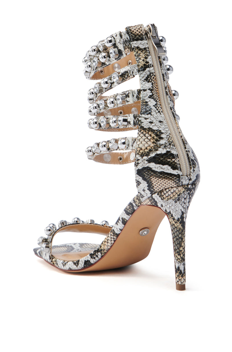 The AZALEA WANG HARVEY MULTI SNAKE SANDAL features vegan leather with a striking snakeskin pattern, silver-studded straps, back zip closure, slender stiletto heel, and a tan-colored inner sole for added flair.