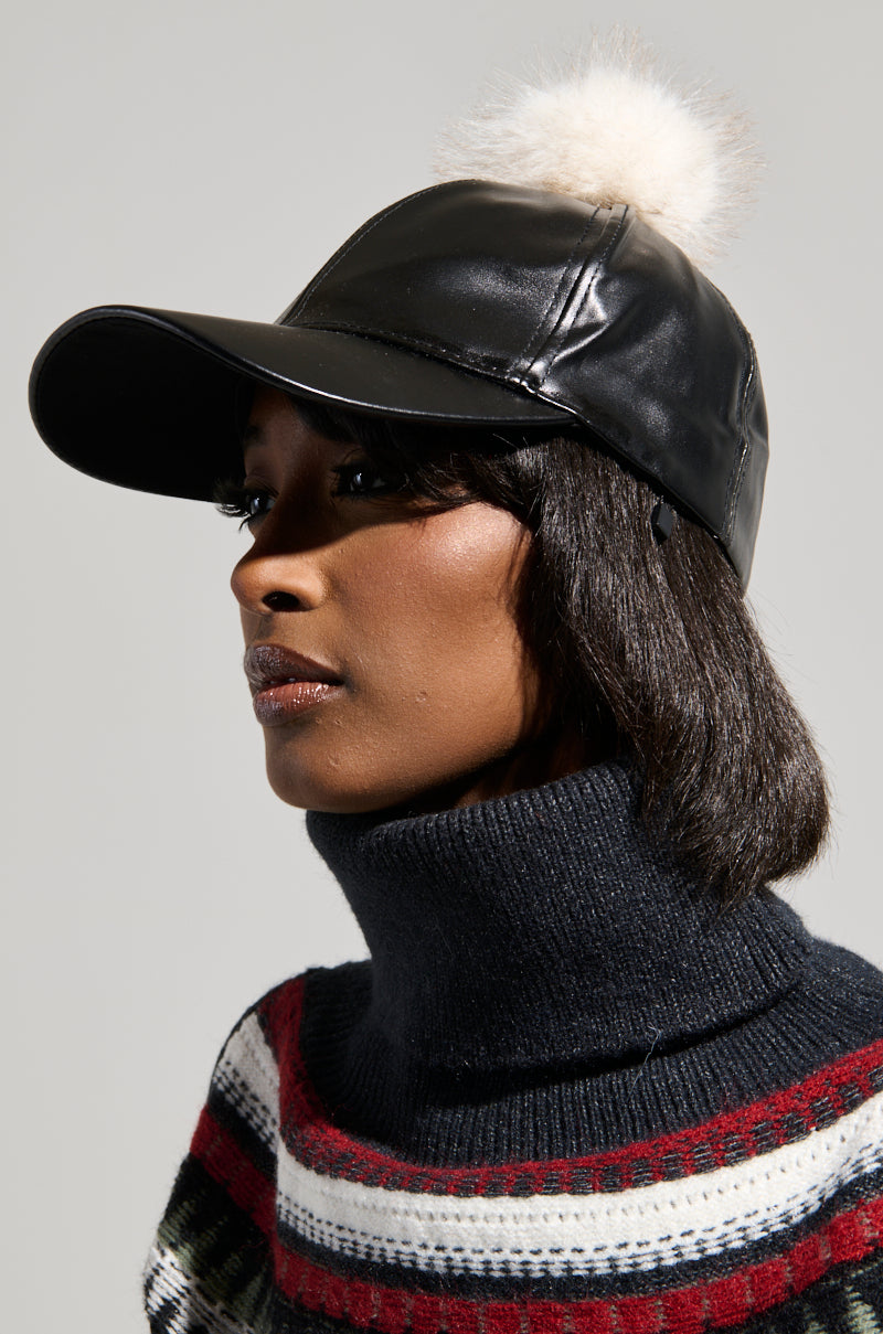 A person wearing the TOASTIE FAUX FUR POM HAT, which is black with a white pom-pom, pairs it with a dark turtleneck sweater featuring red, white, and green stripes. The background is a neutral gray.