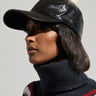 A person wearing the TOASTIE FAUX FUR POM HAT, which is black with a white pom-pom, pairs it with a dark turtleneck sweater featuring red, white, and green stripes. The background is a neutral gray.