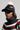 A person wearing the TOASTIE FAUX FUR POM HAT, a black leather baseball cap with an adjustable strap and a white faux fur pom-pom, sits atop their knitted turtleneck sweater featuring a lively red, white, and green pattern. The background is a neutral gray.