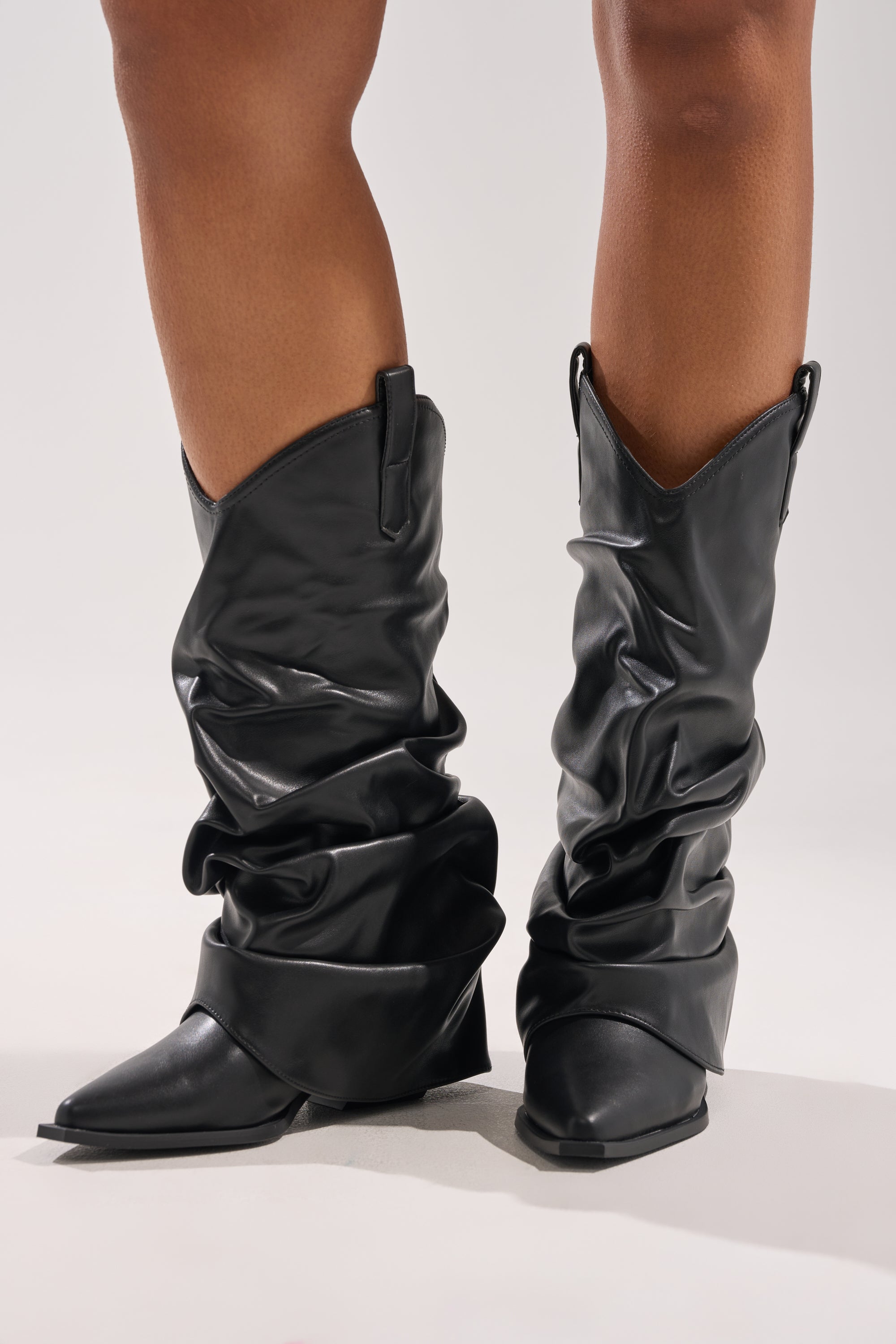 Someone stands on a light-colored surface wearing the AZALEA WANG HAWKSTON BLACK PU RUCHED WESTERN BOOTIE, featuring a ruched mid-calf design, pointed toes, and angled heels.