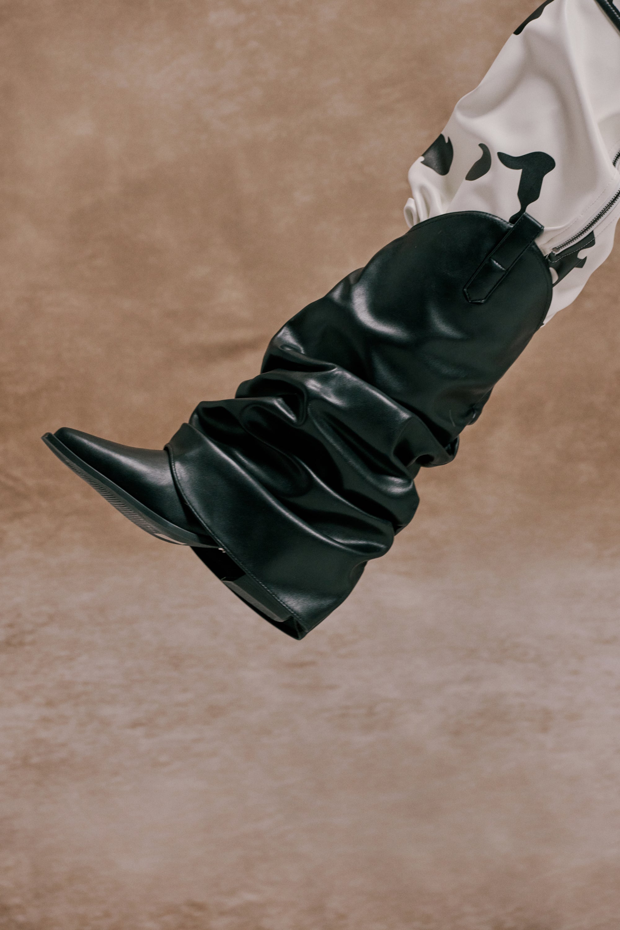 A person wears the AZALEA WANG HAWKSTON BLACK PU RUCHED WESTERN BOOTIE and white pants with black cow-print spots, stretching their leg against a textured brown background. 0