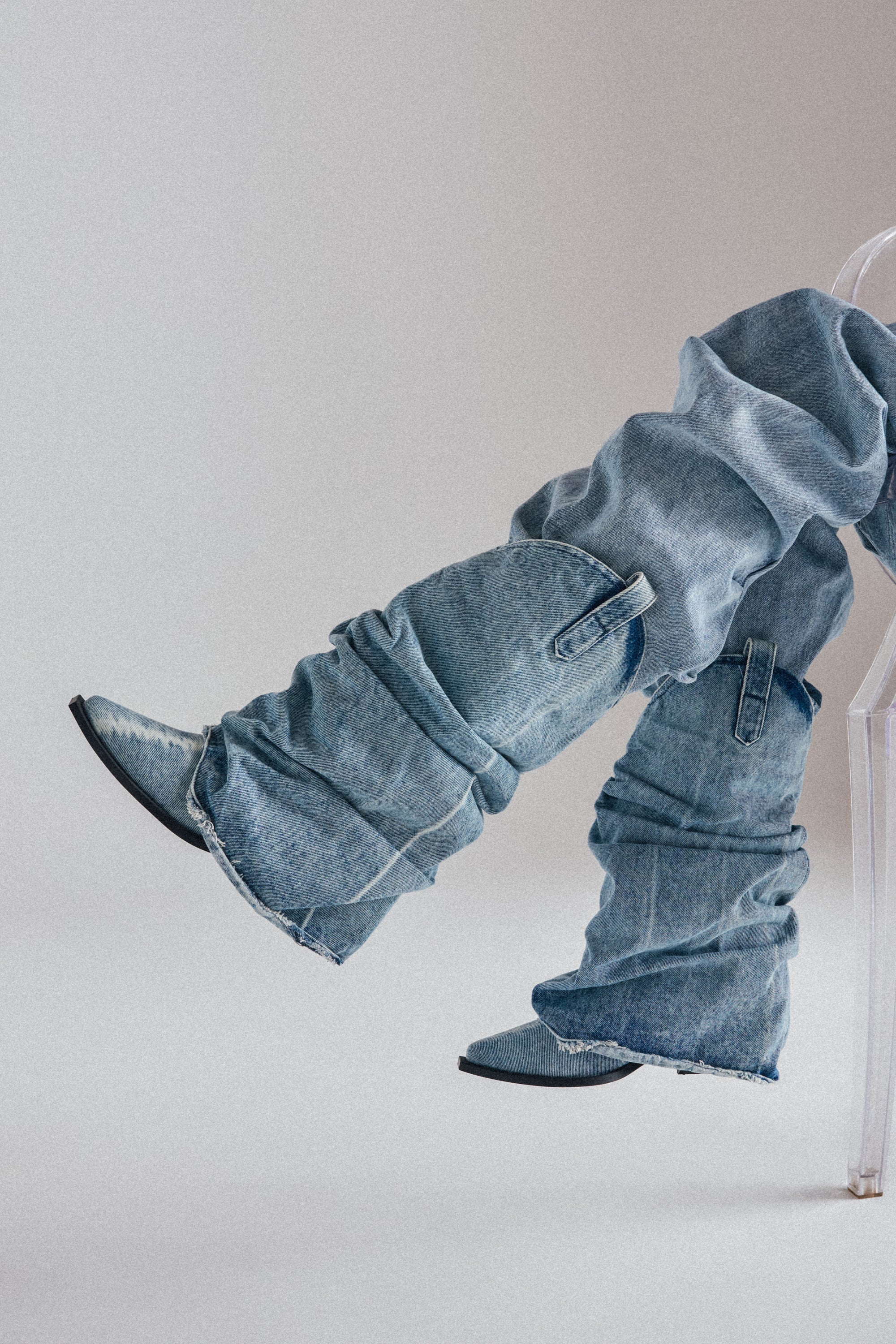 A person in loose, stacked light-wash denim jeans and AZALEA WANG HAWSTON DENIM WESTERN FOLD-OVER BOOTS with a chunky heel sits on a clear chair, legs crossed, against a plain background. 1