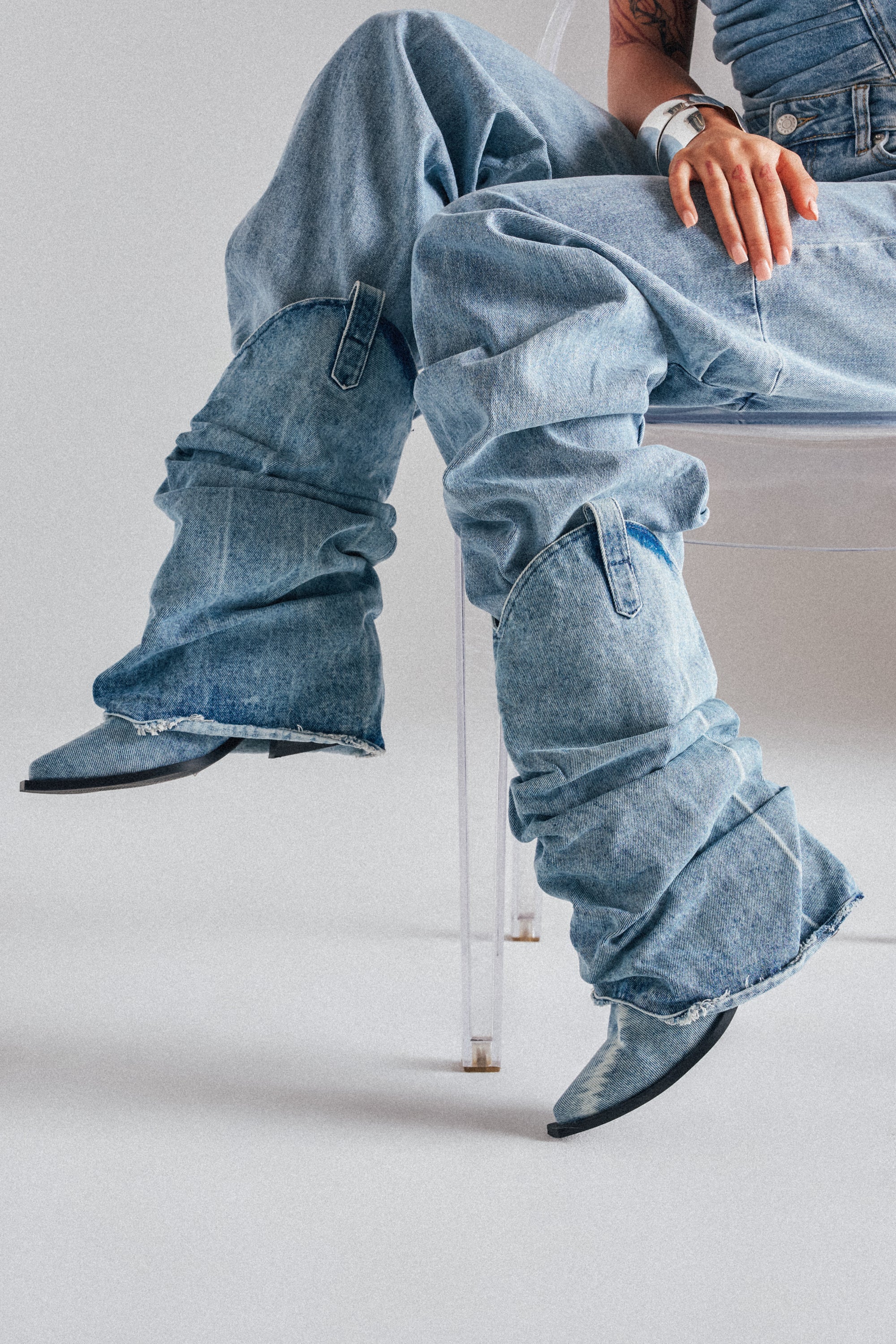 A person seated on a clear chair wears loose light blue jeans over AZALEA WANG HAWSTON DENIM WESTERN FOLD-OVER BOOTS with chunky heels, one hand on their knee and metallic bracelets showing. 3