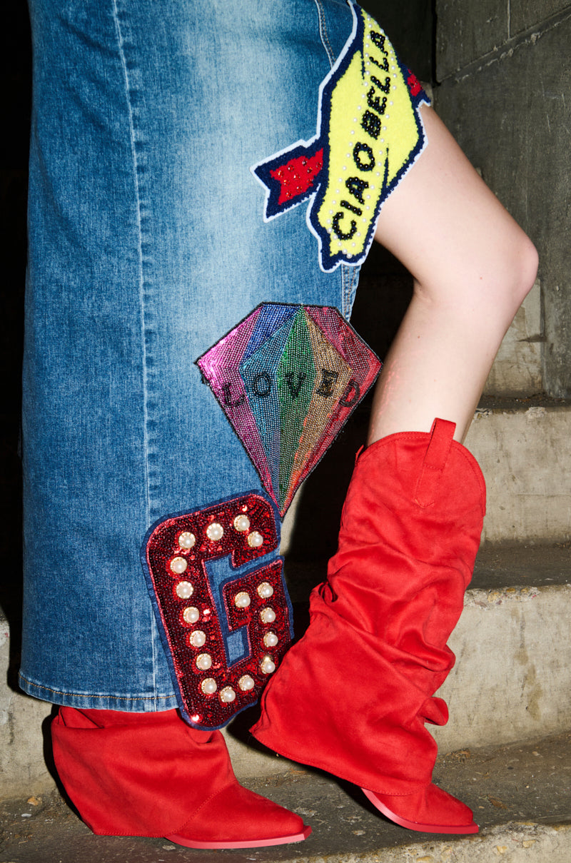 A person in a denim skirt with diamond, "LOVE," and "G" patches stands on concrete steps wearing the AZALEA WANG HAWKSTON RED RUCHED WESTERN BOOTIE with a chunky heel.