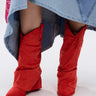 A person wears the AZALEA WANG HAWKSTON RED RUCHED WESTERN BOOTIE with a faux suede upper and chunky heel, paired with a blue patchwork denim skirt featuring a pink bandana detail at the hem, set against a plain light background.