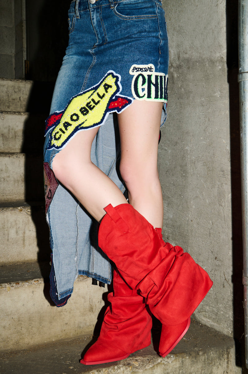 A person stands on concrete steps in a denim skirt with colorful patches, including "CIAO BELLA," and bold AZALEA WANG HAWKSTON RED RUCHED WESTERN BOOTIES. The photo is cropped to highlight the standout boots and lower half of the body.