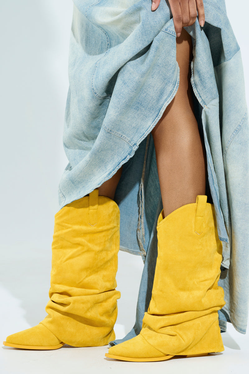 Someone wearing AZALEA WANG HAWKSTON YELLOW BOOTIE stands with one foot forward. Their hand lifts a blue denim skirt to show part of their leg against a plain light background.
