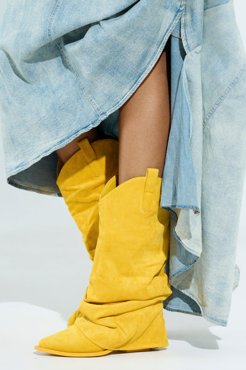 A person wears a flowing light blue denim skirt paired with AZALEA WANG HAWKSTON YELLOW BOOTIE, highlighting the contrast between the textured boots and the smooth denim fabric. The focus is on their legs, emphasizing this striking combination.