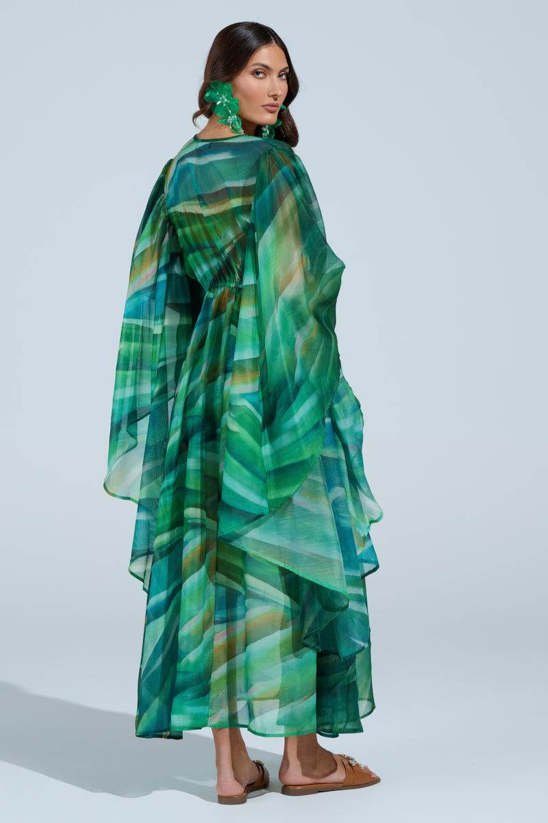 A woman stands slightly to the side in the ON FIRE SET, featuring a flowing multicolored green dress with an abstract wave print. Her dark hair is styled down, enhanced by large green earrings and brown sandals against a light blue background. 10
