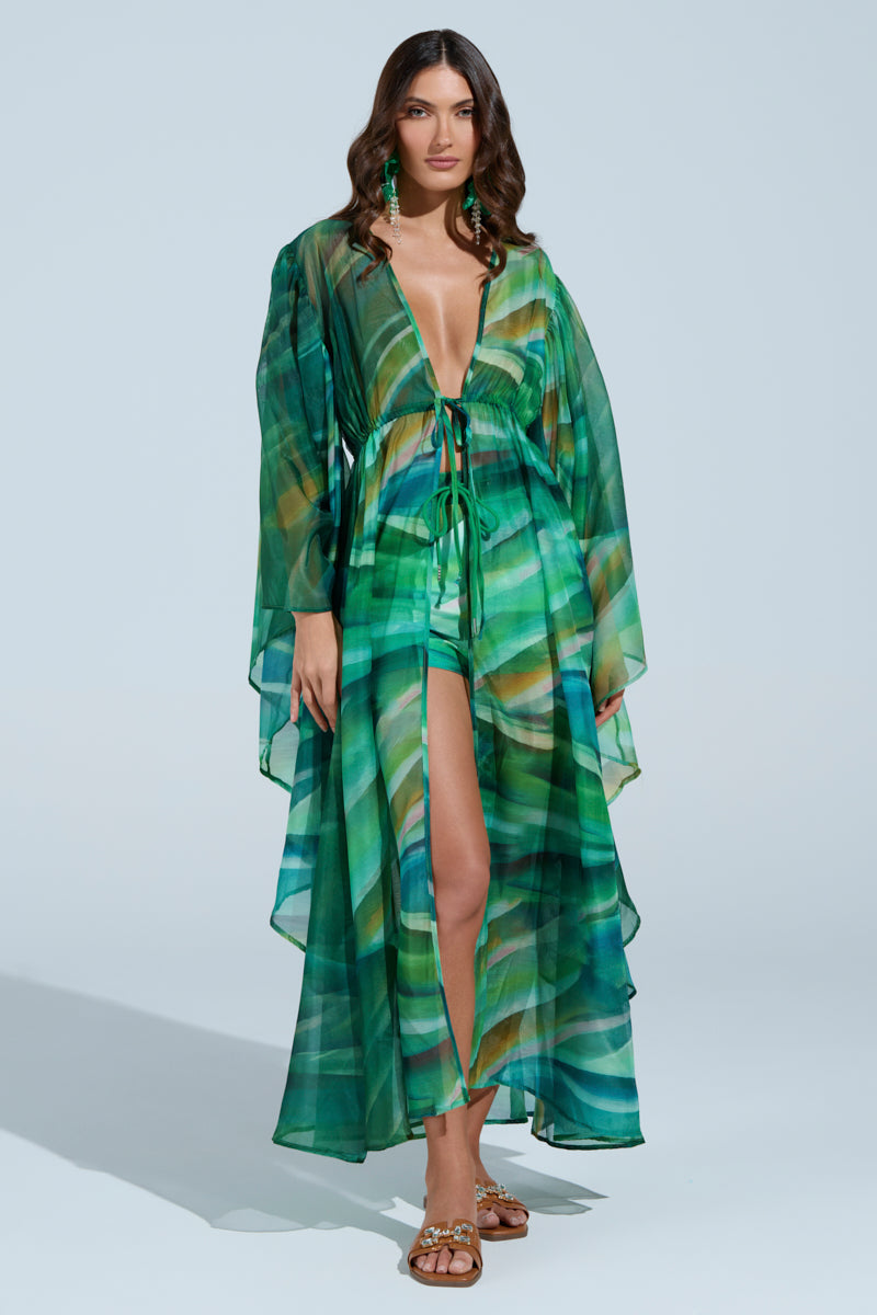 A woman confidently stands in the ON FIRE SET, a robe-inspired top featuring an abstract wavy print, with a plunging neckline and high slit. Her long wavy hair and sandals complement the look against a plain light blue background. 1