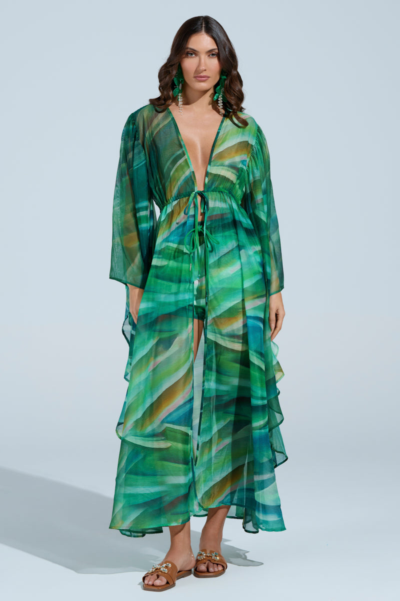 A woman poses against a plain backdrop in the ON FIRE SET—a green, flowing, semi-transparent dress with an abstract leaf-like wavy print. It boasts a deep V-neck and waist tie, perfectly complemented by her brown sandals for an elegant ensemble. 3