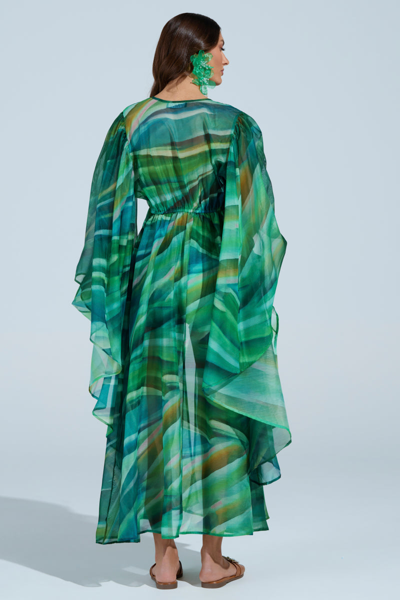 A person models the ON FIRE SET, featuring a flowing dress with abstract green and blue wavy print and sheer draped sleeves. The cinched waist and full-length skirt complement large green earrings and sandals against a plain light blue backdrop. 5