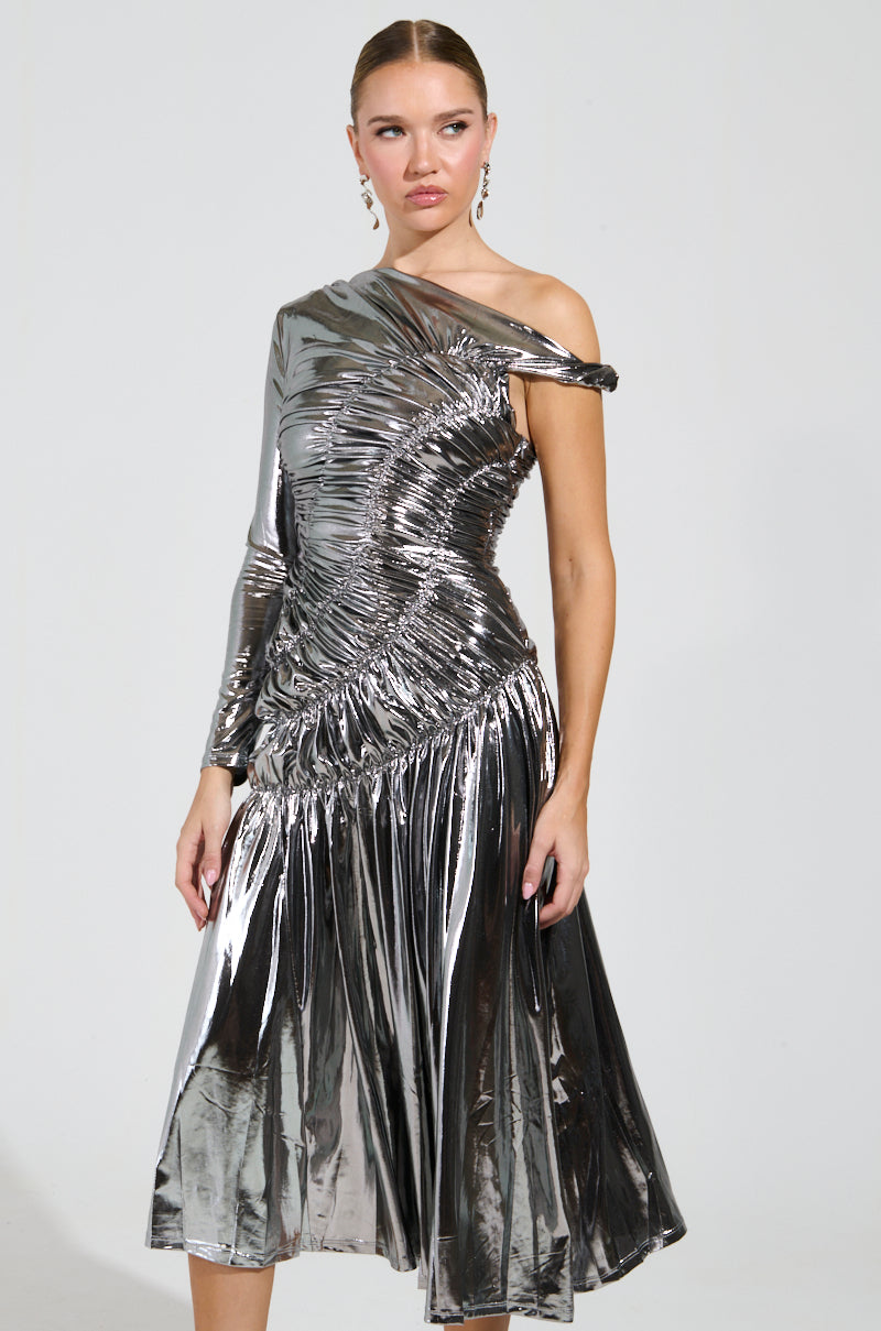 Against a plain background, an individual stands in the TURN BACK TIME SILVER METALLIC MIDI DRESS, which features a striking asymmetrical neckline with detailed ruching. The dress highlights a fitted bodice and seamlessly flows into its elegant skirt. Their hair is pulled back, showcasing the sophisticated earrings that perfect the ensemble.
