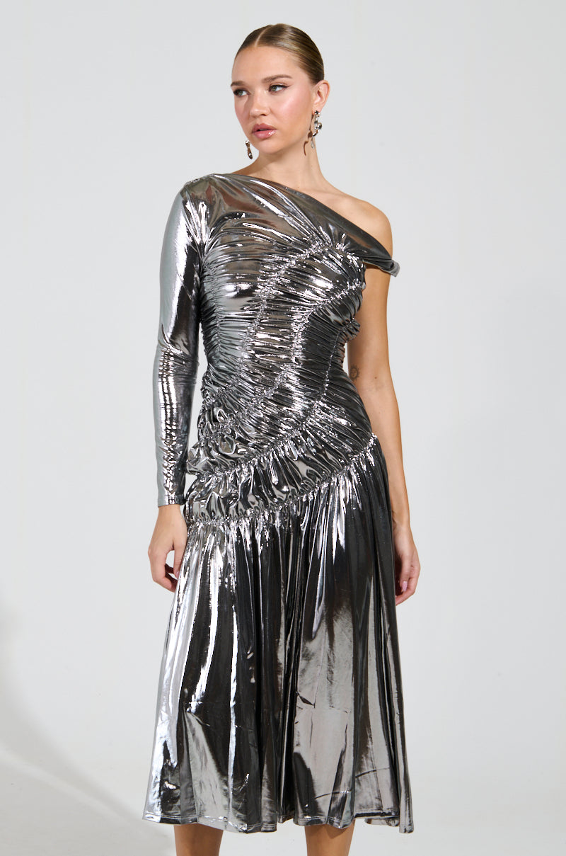 A woman poses in the TURN BACK TIME SILVER METALLIC MIDI DRESS, showcasing its ruched bodice along with its asymmetrical neckline and hemline. She accessorizes with elegant earrings and has her hair styled in an updo, set against a plain background.