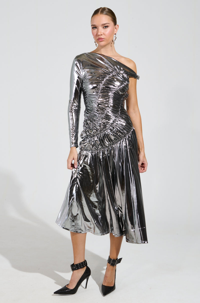 A person is wearing the TURN BACK TIME SILVER METALLIC MIDI DRESS, which features a ruched bodice and an asymmetrical hemline. They complete the look with sleek black high-heeled shoes against a plain, light-colored background.