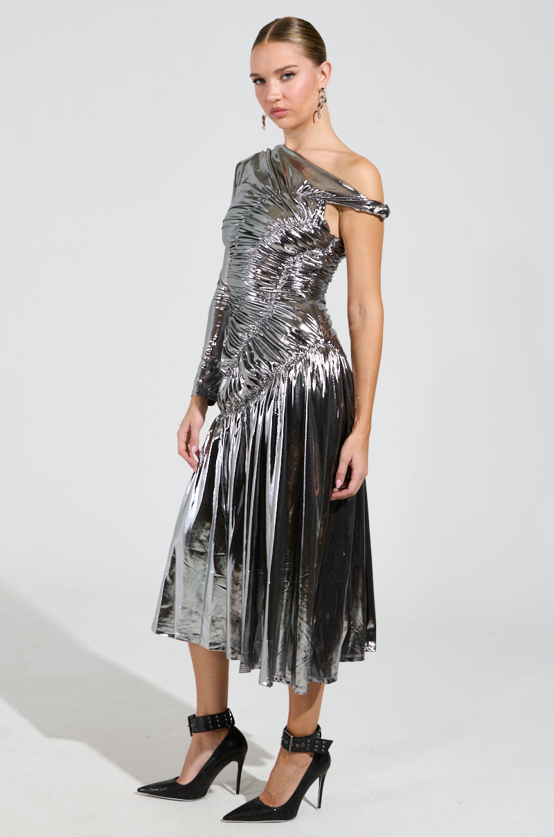 A woman poses elegantly in the TURN BACK TIME SILVER METALLIC MIDI DRESS, which boasts an asymmetrical neckline and pleated skirt. She finishes her look with black pointed heels and large hoop earrings, standing against a plain light background.