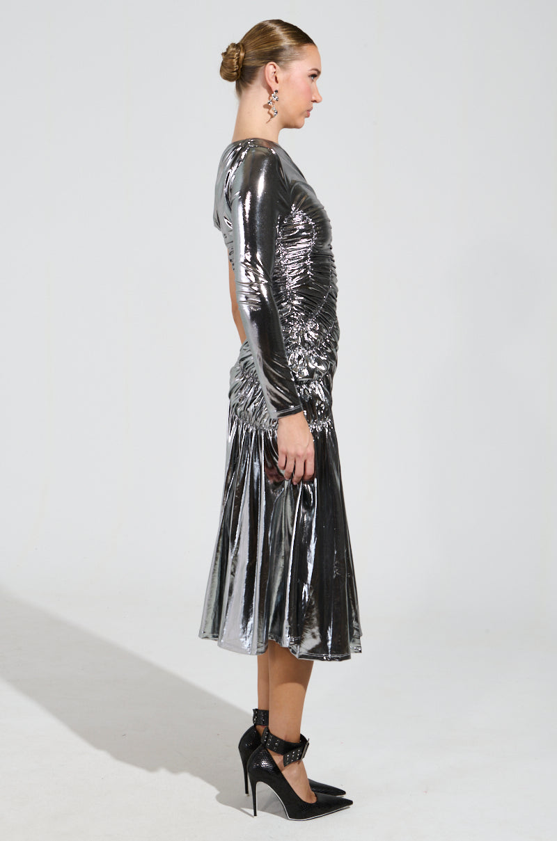 A person dressed in the TURN BACK TIME SILVER METALLIC MIDI DRESS, which features long sleeves and a ruched bodice that adds texture, stands in profile against a plain background. Black high heels and a neat updo complete their look, with dangling earrings visible.