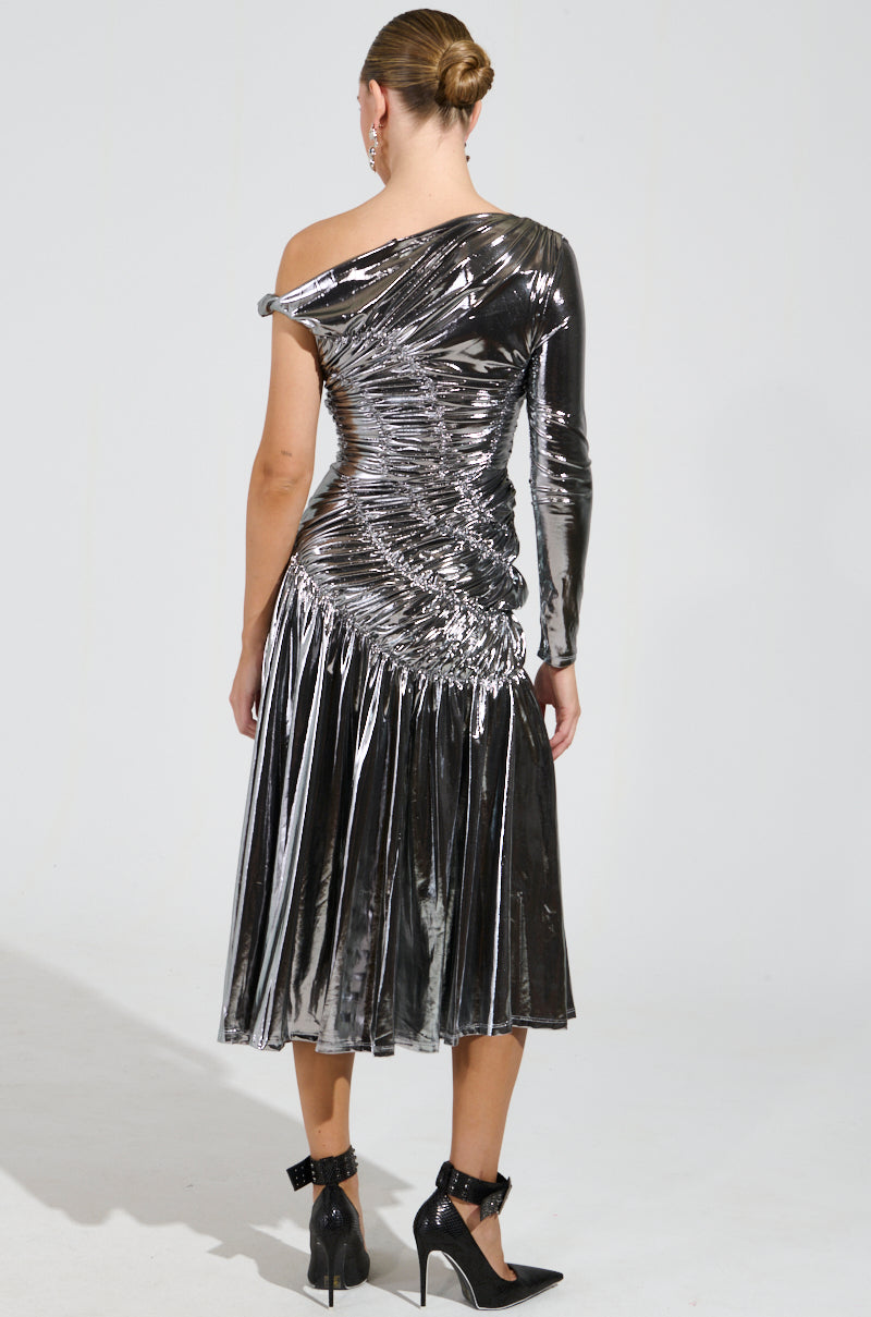 Dressed in the TURN BACK TIME SILVER METALLIC MIDI DRESS, a person stands with their back to the camera, showcasing its asymmetrical neckline. The ruched bodice lends texture to the outfit, which falls gracefully to mid-calf. High-heeled shoes complete the ensemble, while their hair is elegantly styled in a bun.