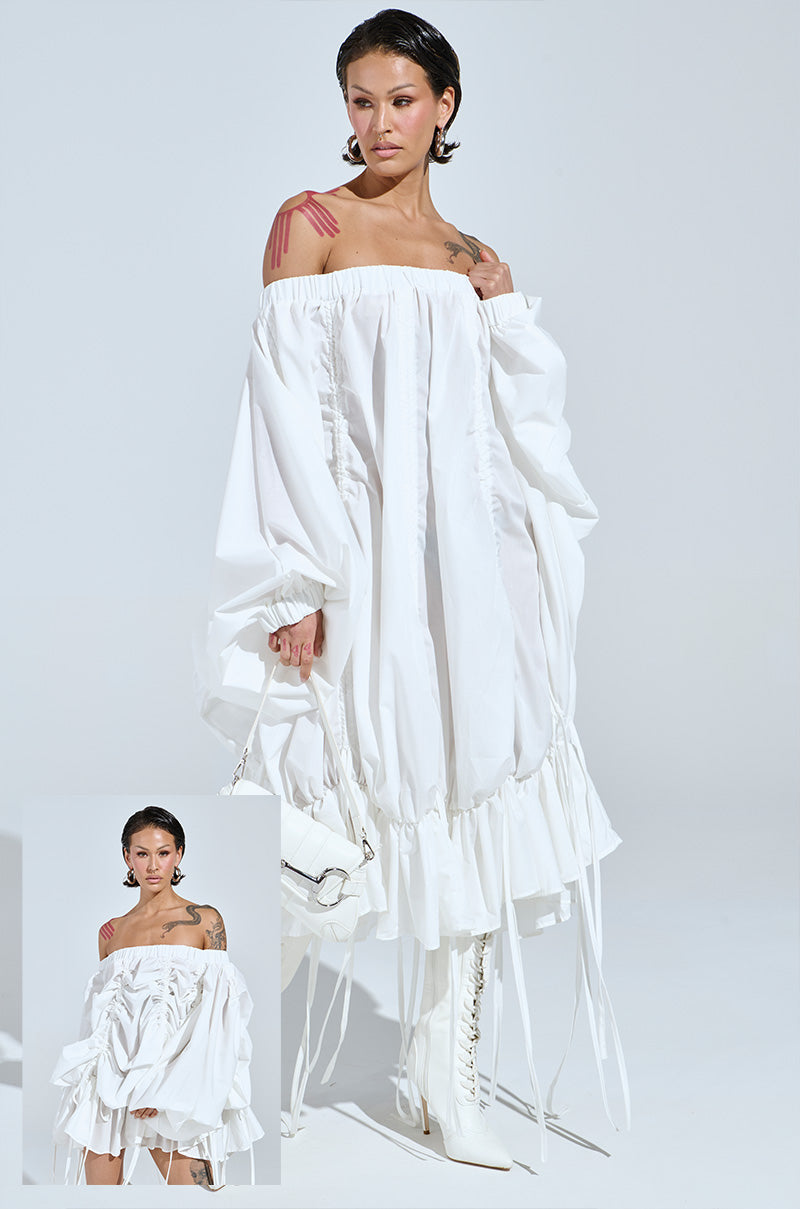 Person in the FREE AS CAN BE MINI DRESS IN WHITE, featuring off-the-shoulder and ruched design with long sleeves, holding a white bag. They stand in knee-high boots with slicked-back hair and visible tattoos. Inset offers a closer look at the chic ensemble.