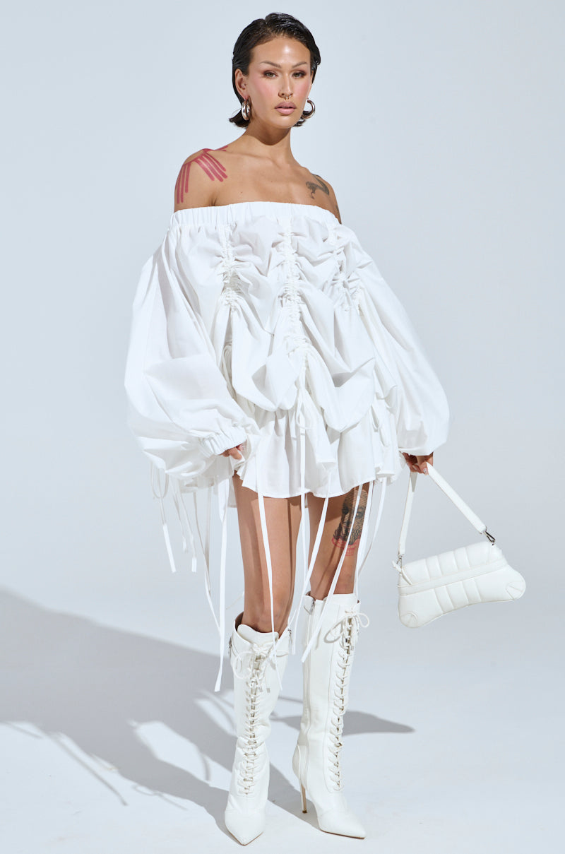 A person poses against a plain backdrop in the FREE AS CAN BE MINI DRESS IN WHITE, showcasing its off-the-shoulder style with ruched detailing and long ties. They accessorize with a stylish white handbag and knee-high white boots, their tattoos visible on shoulder and chest.