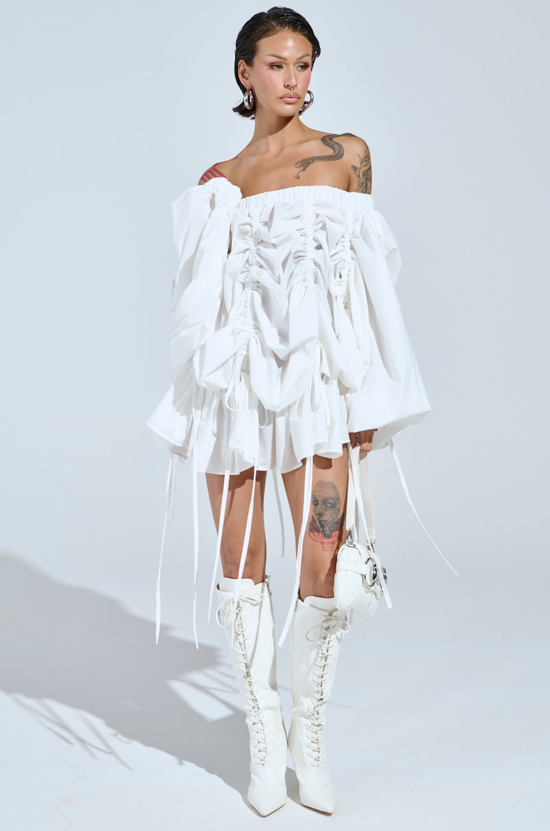 Wearing the chic FREE AS CAN BE MINI DRESS IN WHITE with ruffles and lace-up detailing, they expertly pair it with knee-high lace-up boots. Their tattoos add an edgy flair to the elegant look against a plain backdrop.