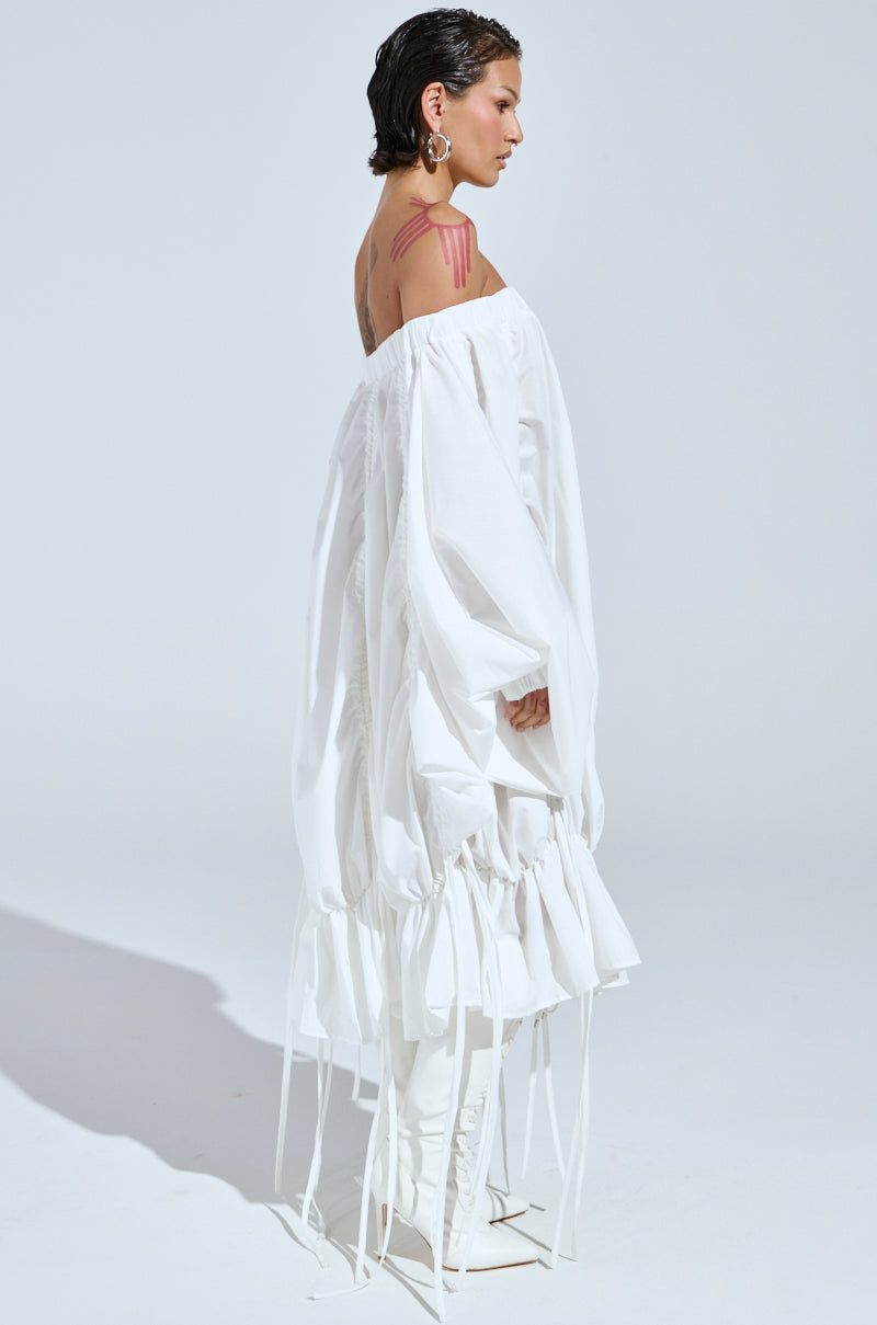 Wearing the "Free As Can Be" mini dress in white, featuring off-the-shoulder, ruched design with long sleeves and gathered details, a person stands in profile. They have slicked-back hair with bold pink makeup lines on their right shoulder and large hoop earrings against a plain white background.