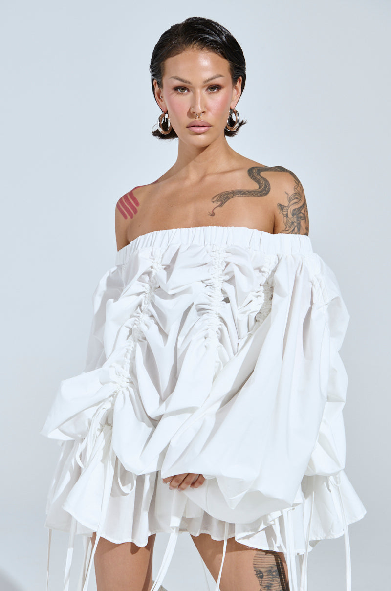 Wearing the FREE AS CAN BE MINI DRESS IN WHITE, a person with tattoos on their arms stands against a plain background. With short dark hair and hoop earrings, they exude confidence and style.